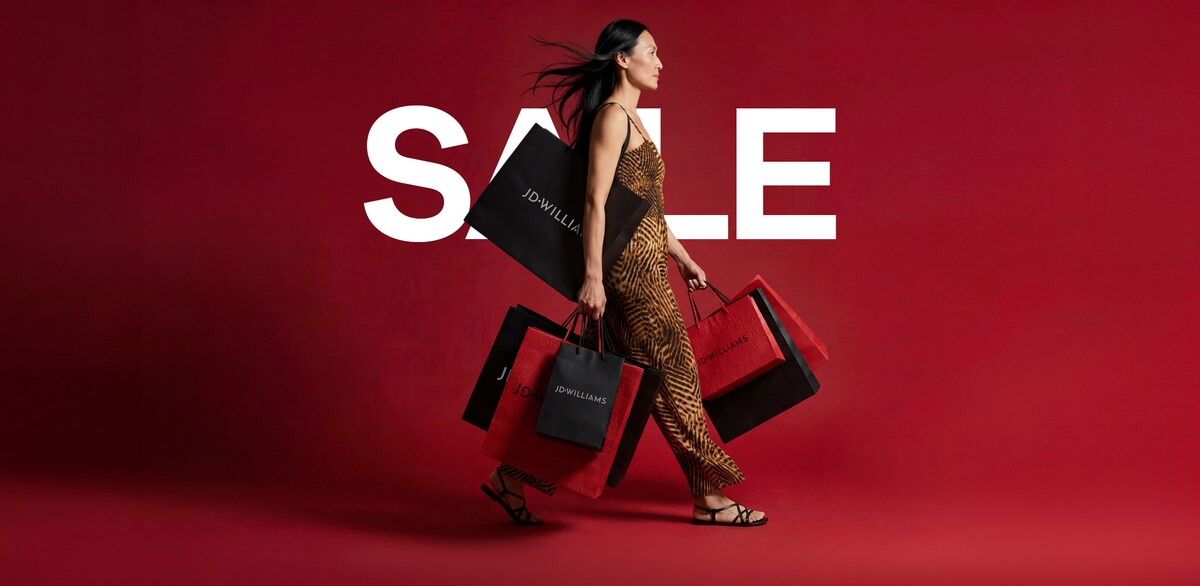 A woman walking with lots of JD Williams shopping bags in front of a red background with the word 'SALE' on it in white