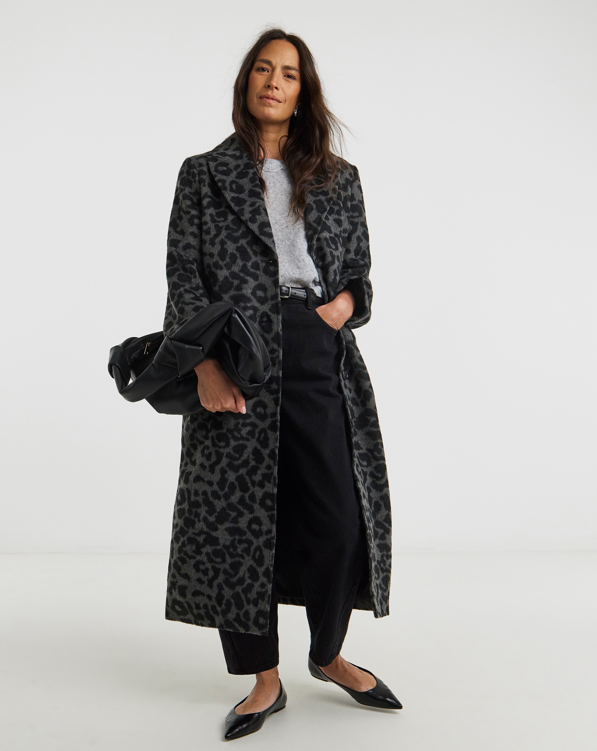 Person wearing a long dark leopard-print coat over a grey top and black trousers, holding a black clutch bag.
