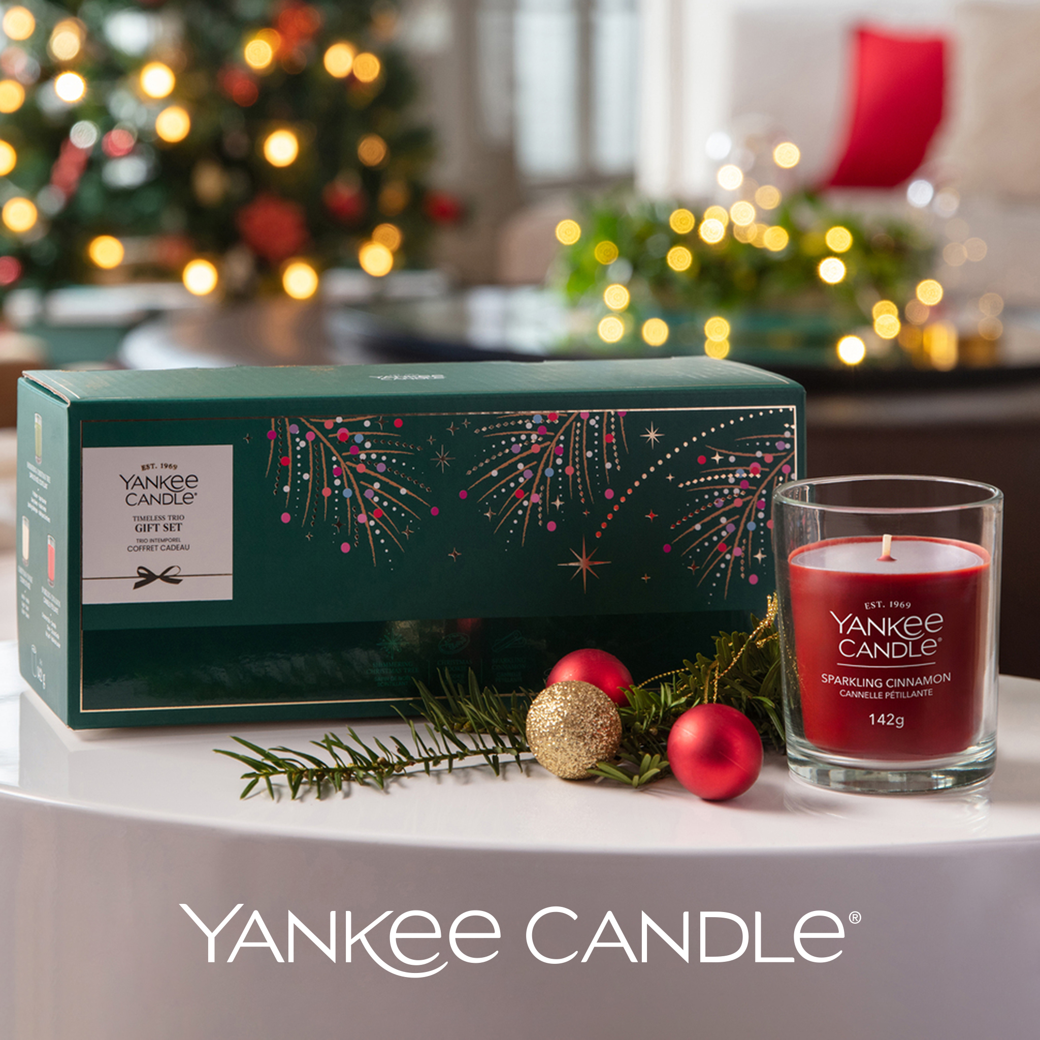 A Yankee Candle Timeless Trio Gift Set box next to a red Sparkling Cinnamon candle, with festive ornaments and a Christmas tree in the background.