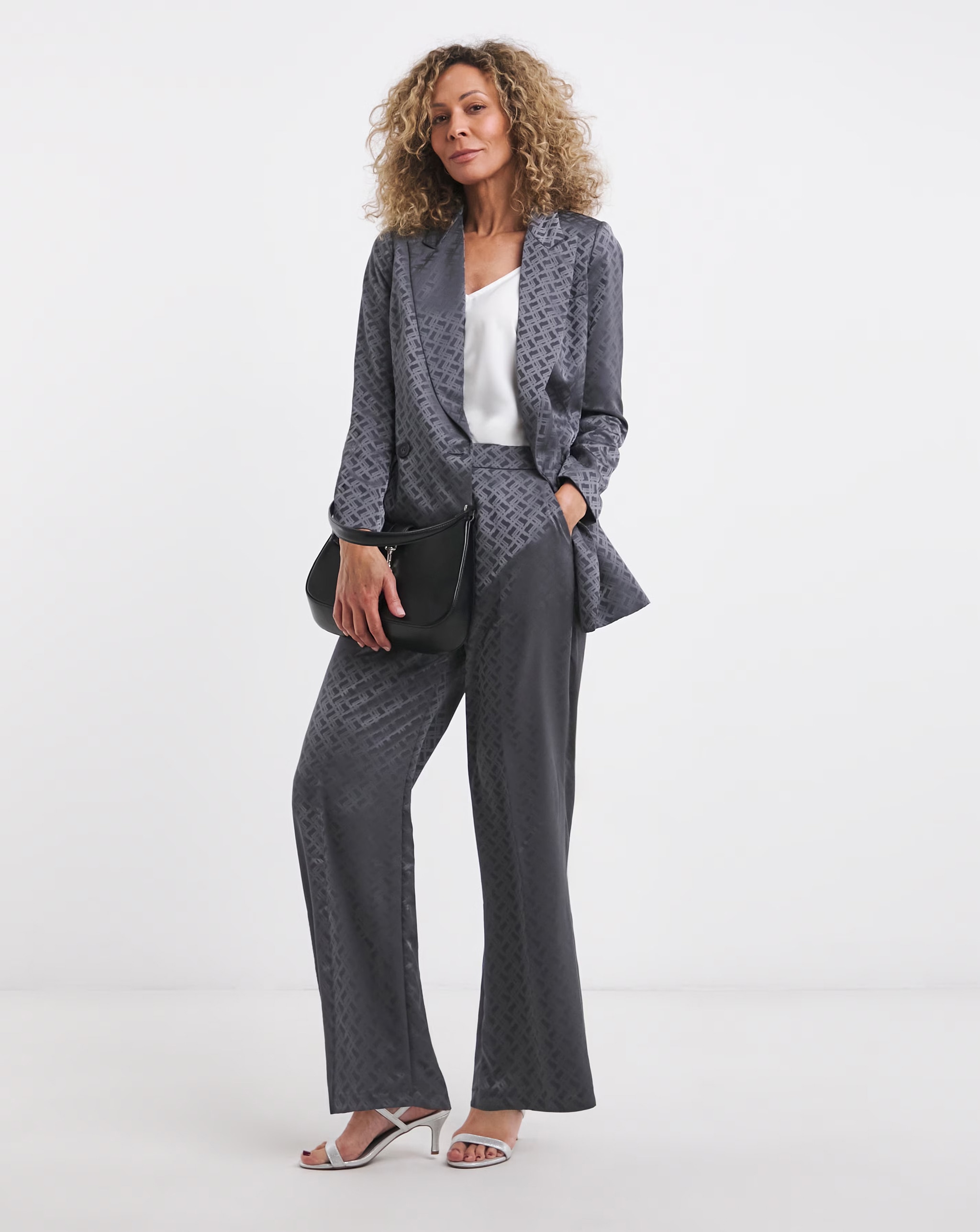 A person stands against a plain white background wearing a matching dark grey patterned blazer and wide‑leg trousers, a white top, and light heeled sandals, holding a black clutch bag.