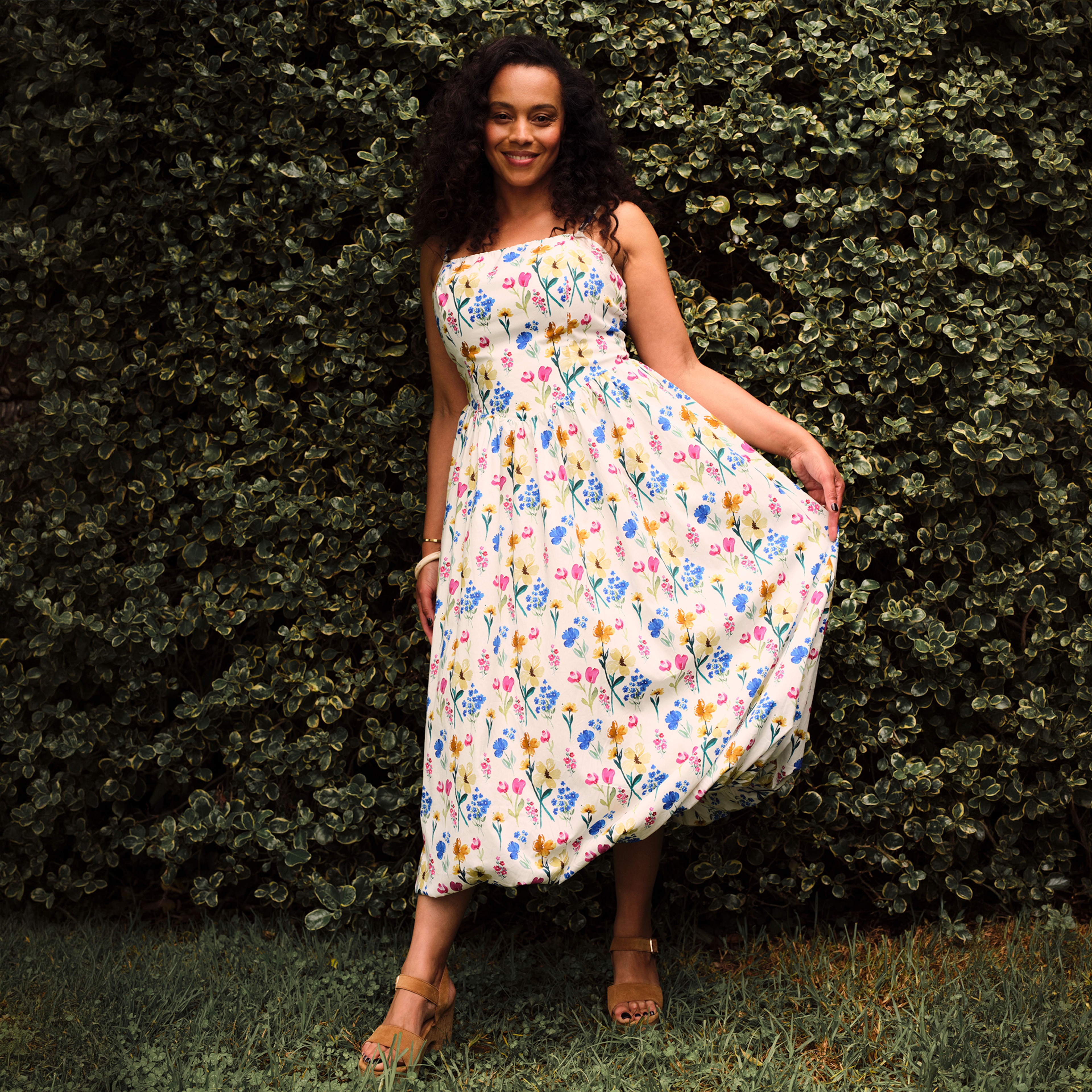 A lady wearing a floral dress in front of a green hedge.