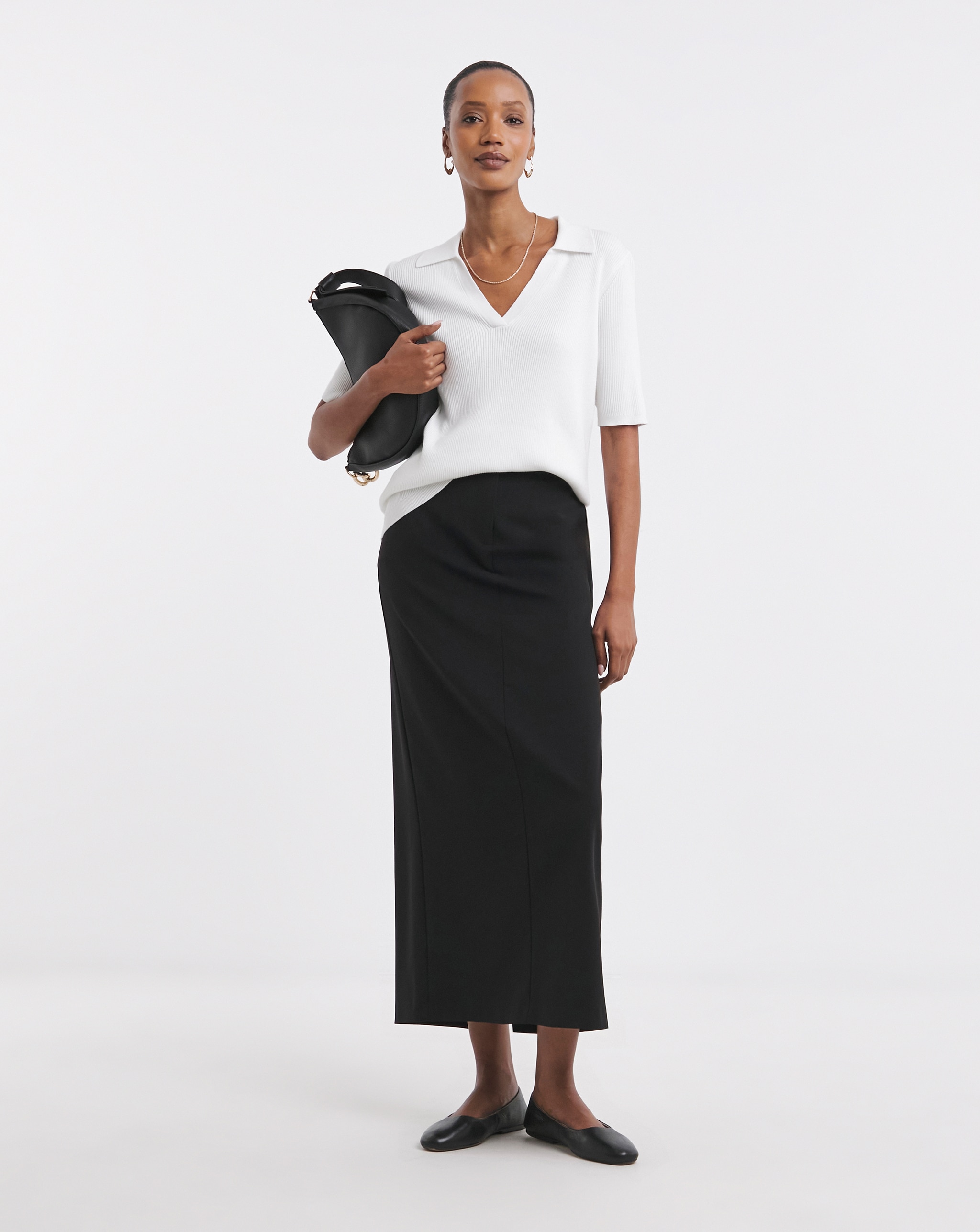 Person wearing a white short‑sleeve top and a long black skirt, holding a black clutch.