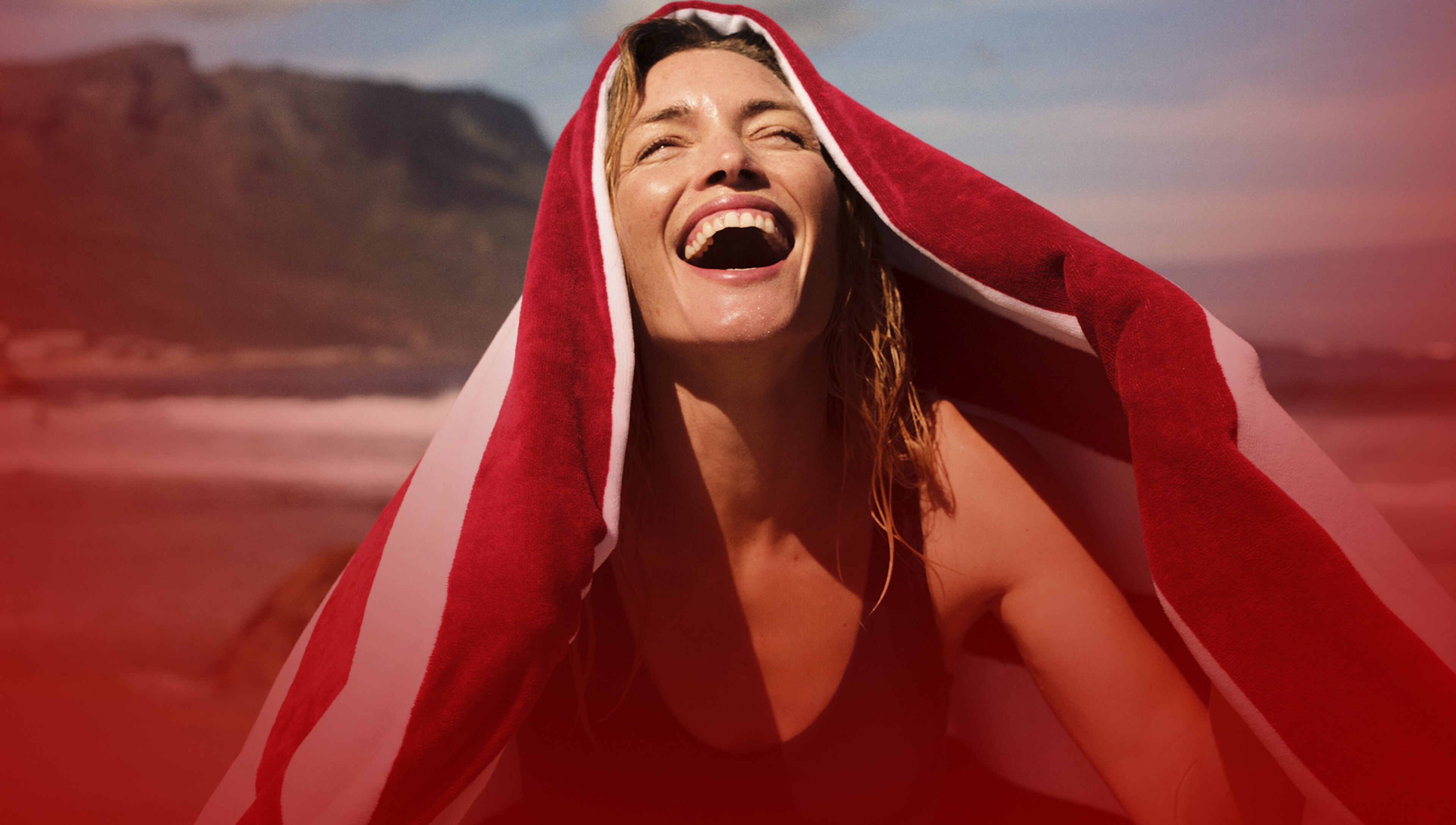 A lady looking up to the sun with her eyes closed smiling with a red and white striped towel over her head.