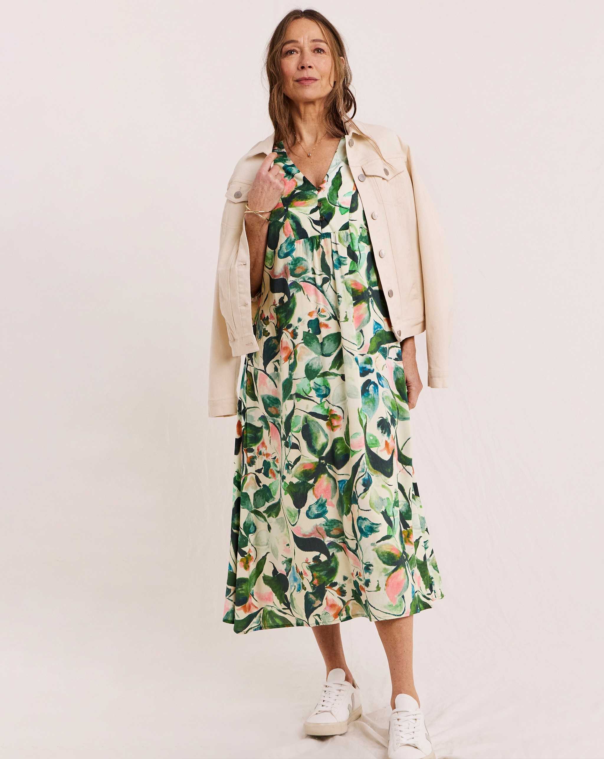 Person wearing a cream denim jacket over a green floral midi dress, styled with white trainers.