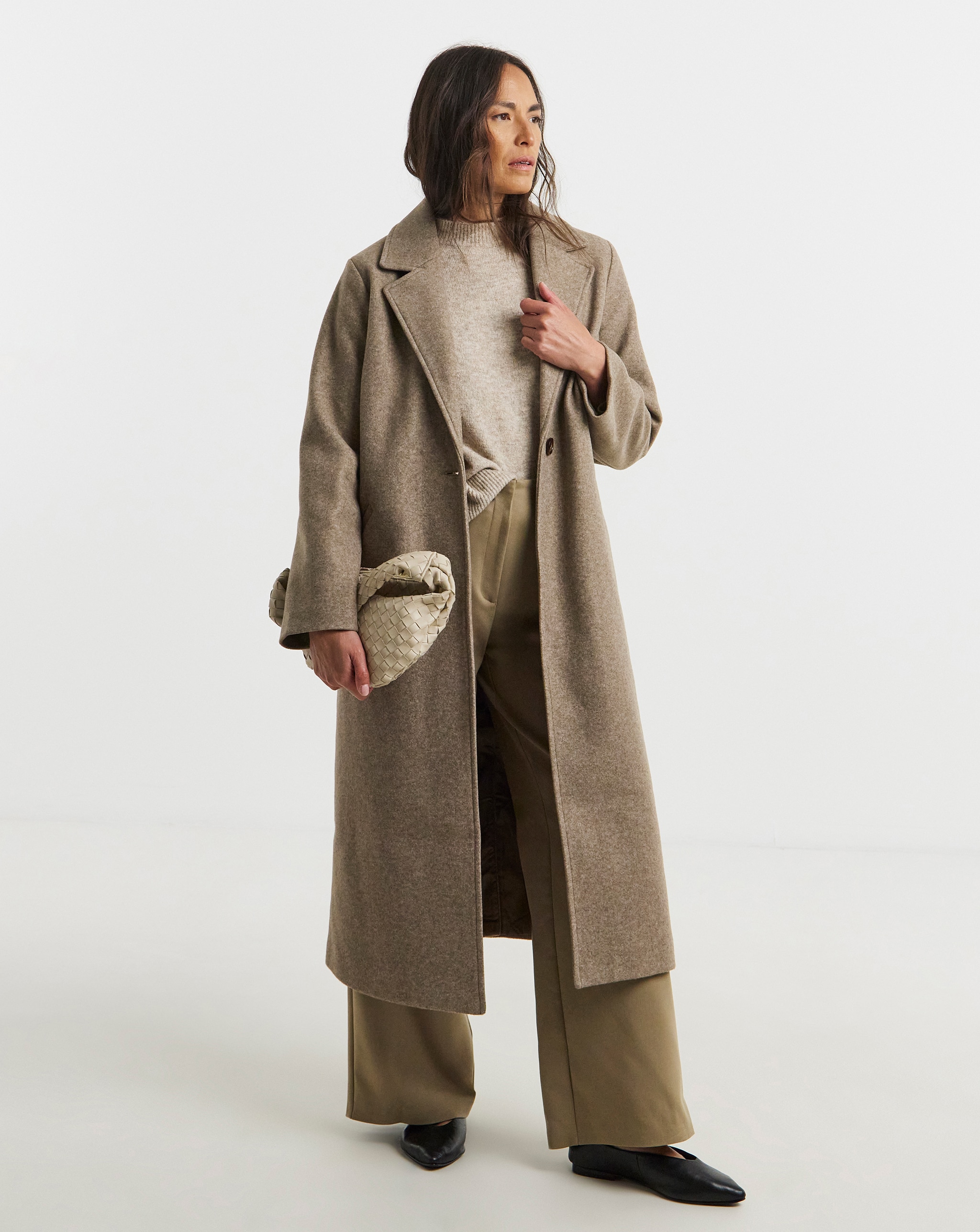 A model standing in a long tan-coloured coat over a jumper and wide‑leg trousers. 