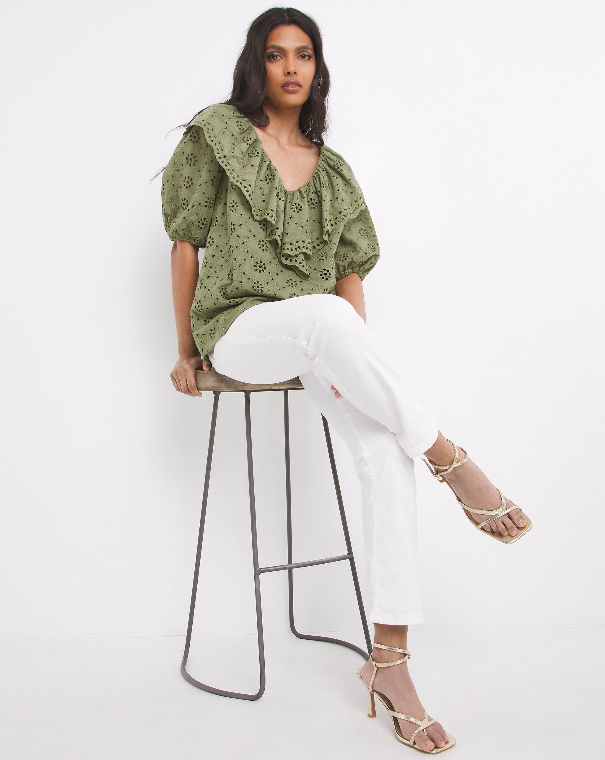 Person seated on a stool, wearing an olive green broderie anglaise blouse with ruffle detailing, white trousers, and strappy metallic heels.