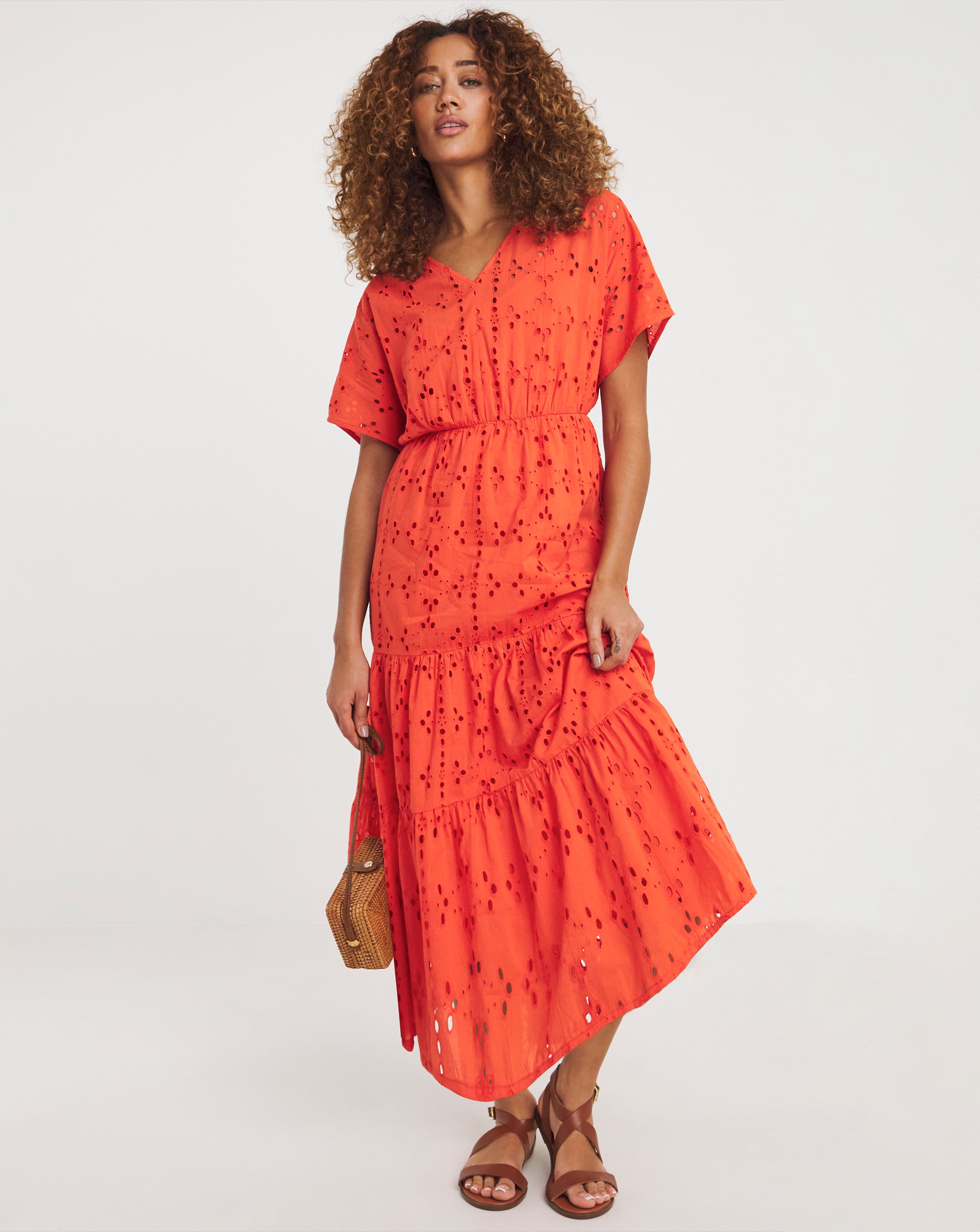 Person wearing a coral broderie anglaise midi dress with short sleeves, brown sandals, and carrying a woven handbag.