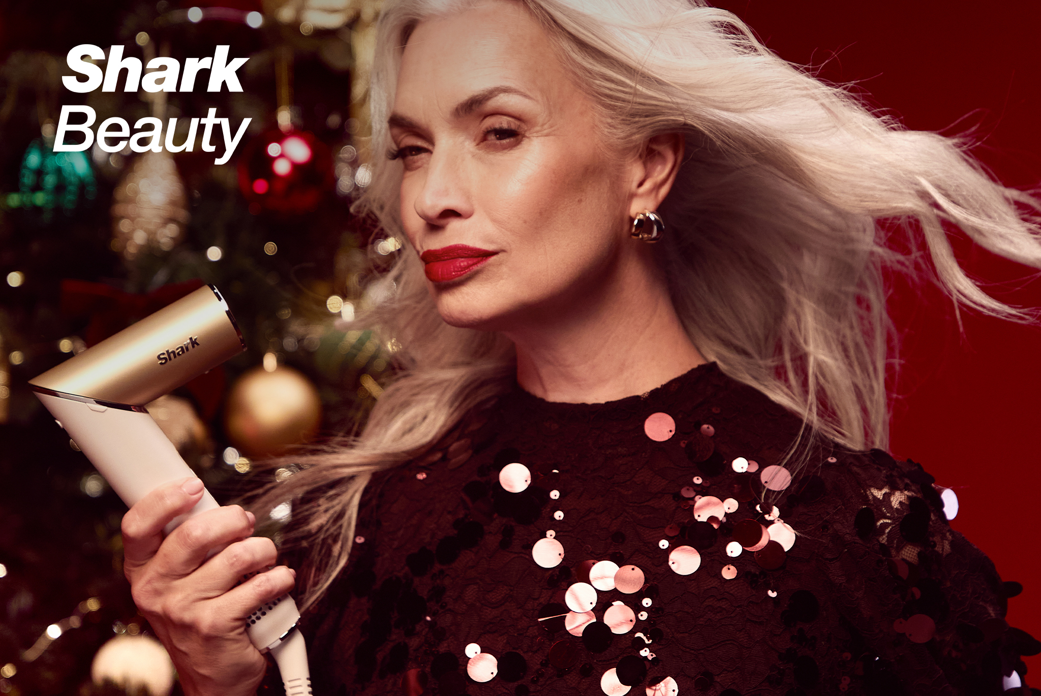 A woman with long, flowing silver-blonde hair and red lipstick is holding a gold and white Shark hair styling tool. She is wearing a black sequined top, with a decorated Christmas tree and festive red background behind her.