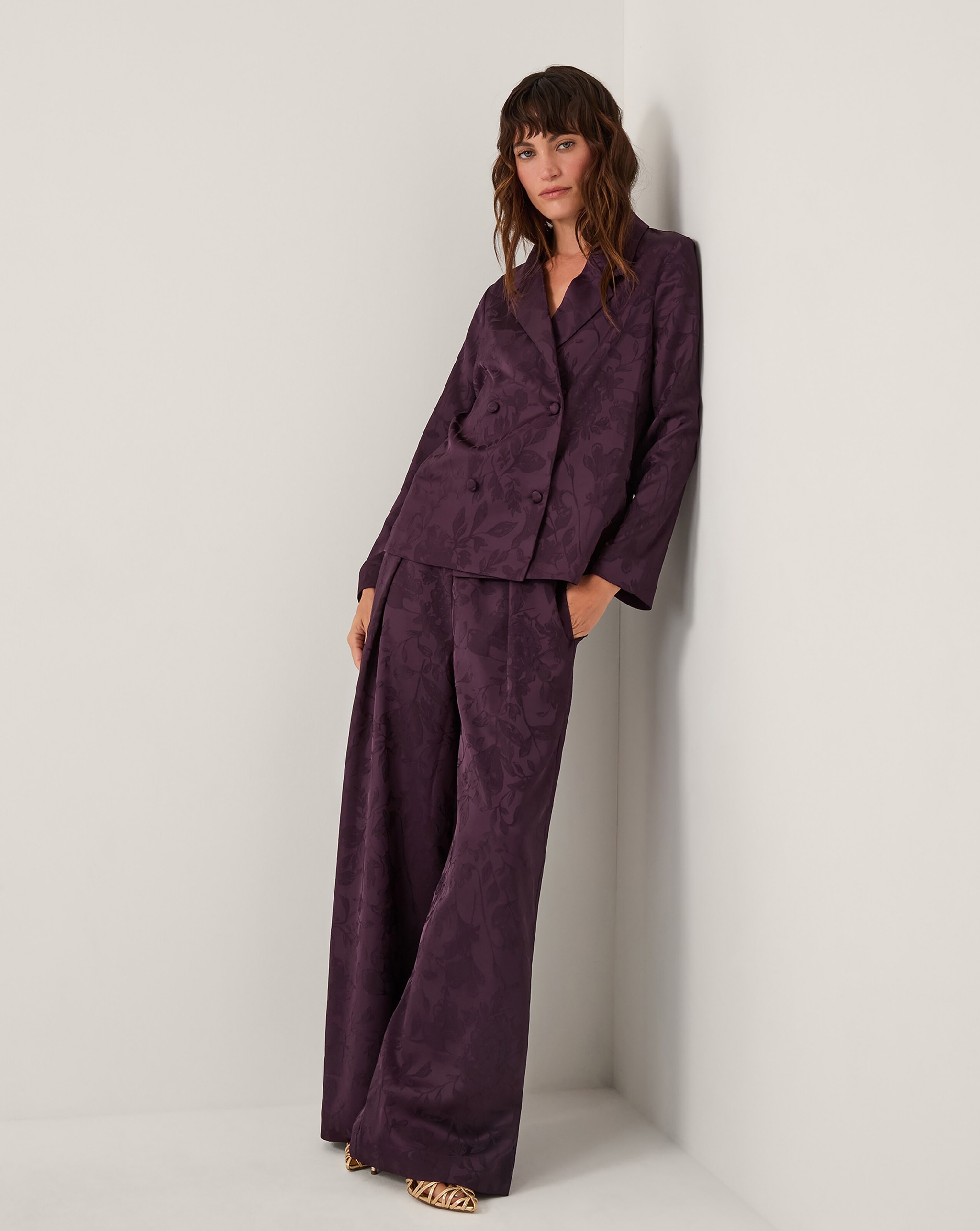 A person leaning against a corner wall wearing a dark purple patterned suit with a double‑breasted jacket and wide‑leg trousers.