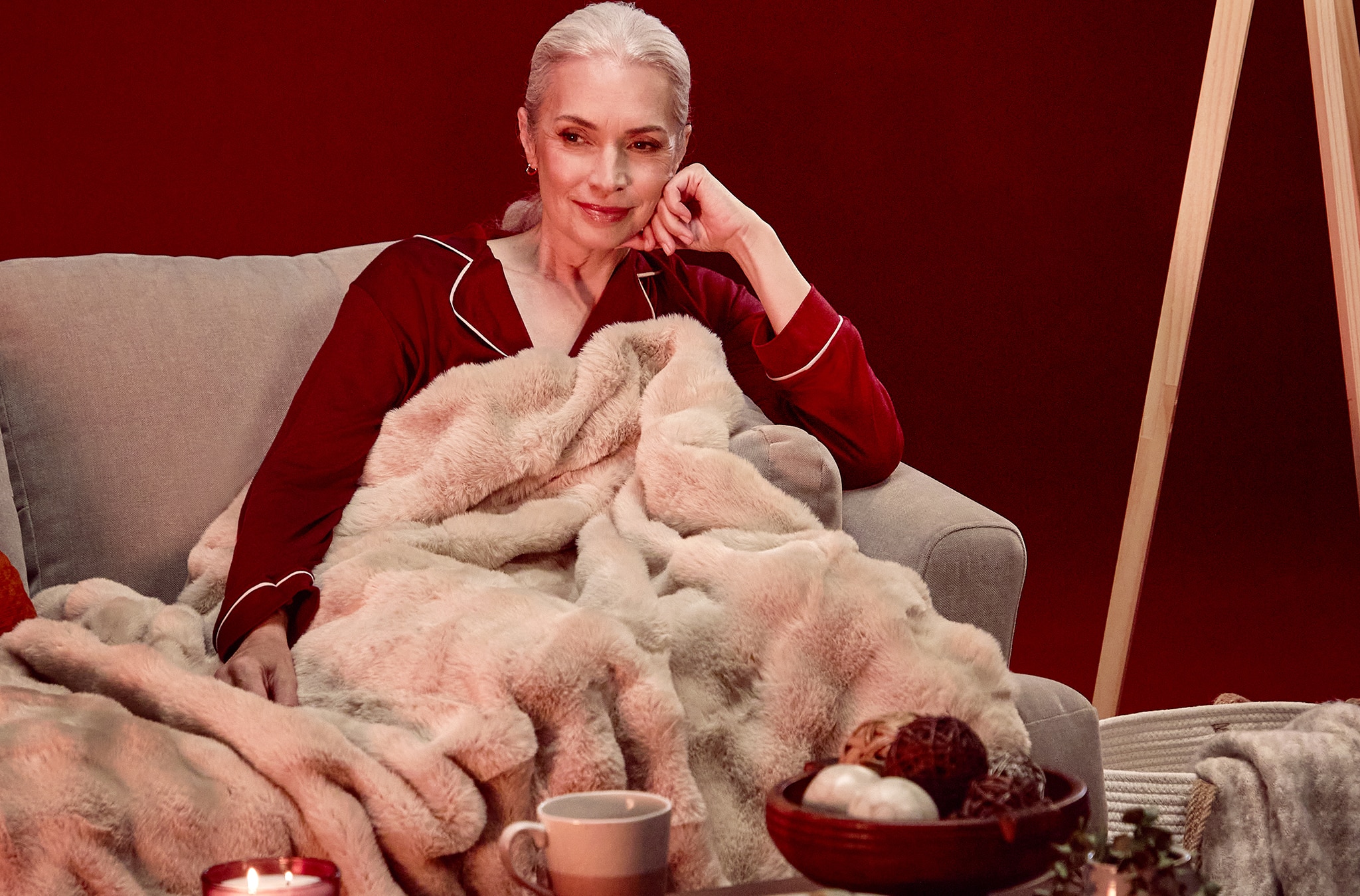 A woman in burgundy pyjamas sat on a sofa under a soft light brown throw.