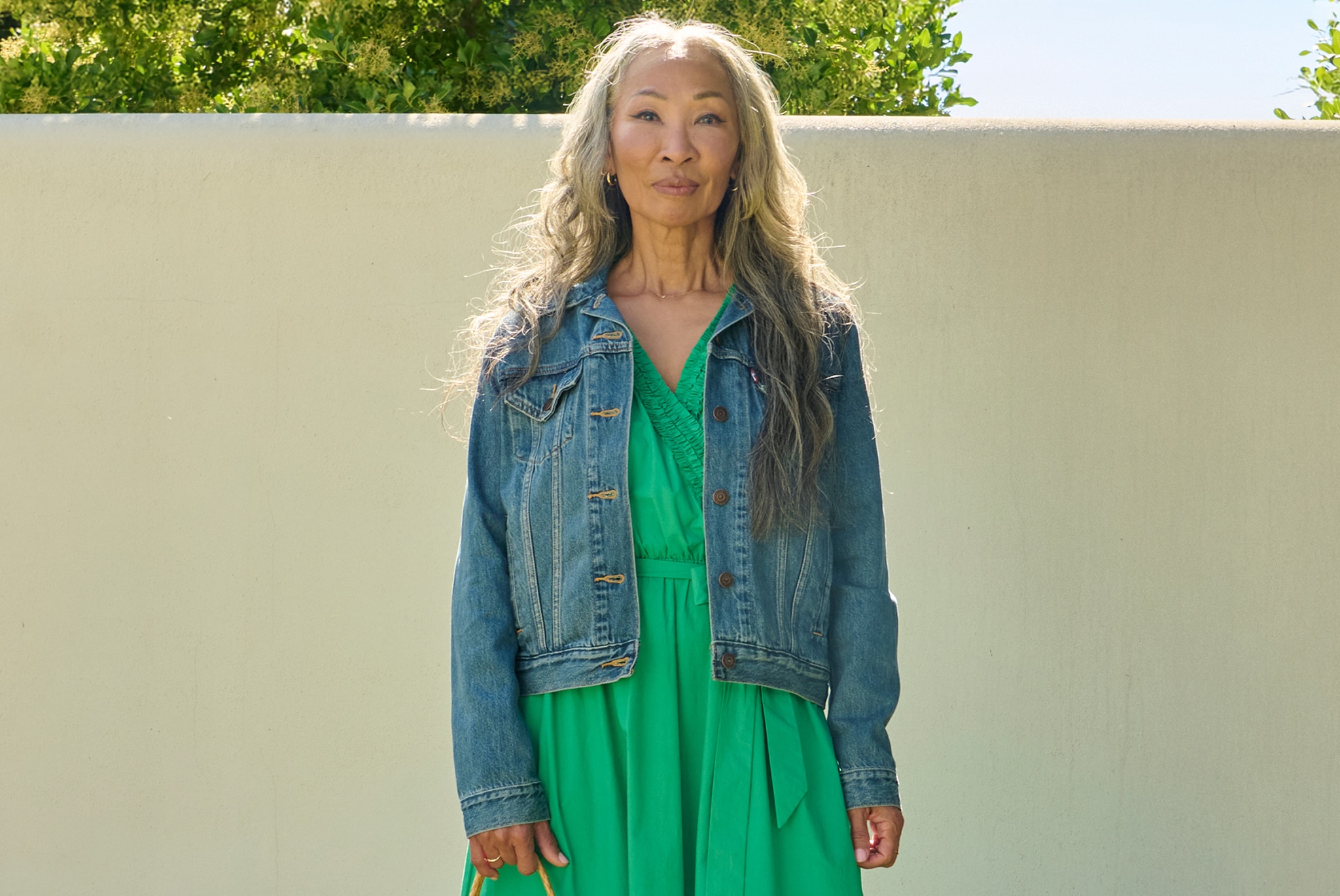 Person wearing a classic blue denim jacket over a bright green wrap-style dress, standing outdoors against a light-coloured wall.