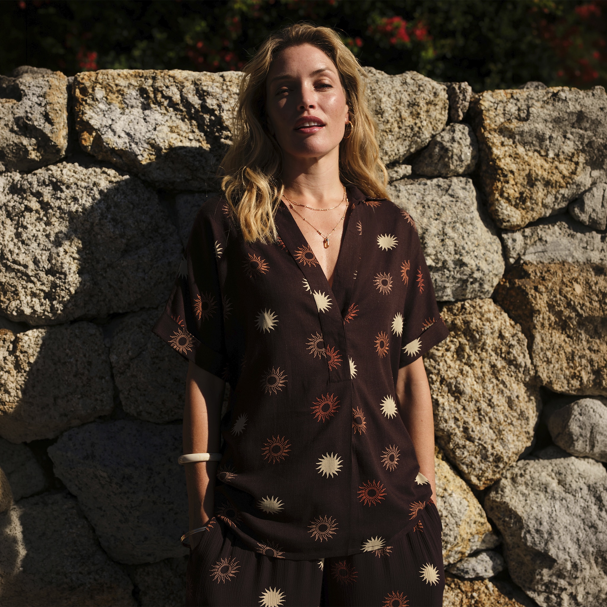 A lady leaning against a wall wearing a brown patterned blouse.