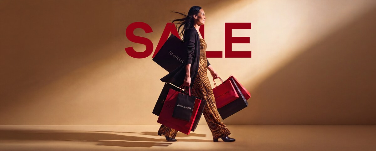 A woman walking from left to right carrying lots of JD Williams branded shopping bags.
