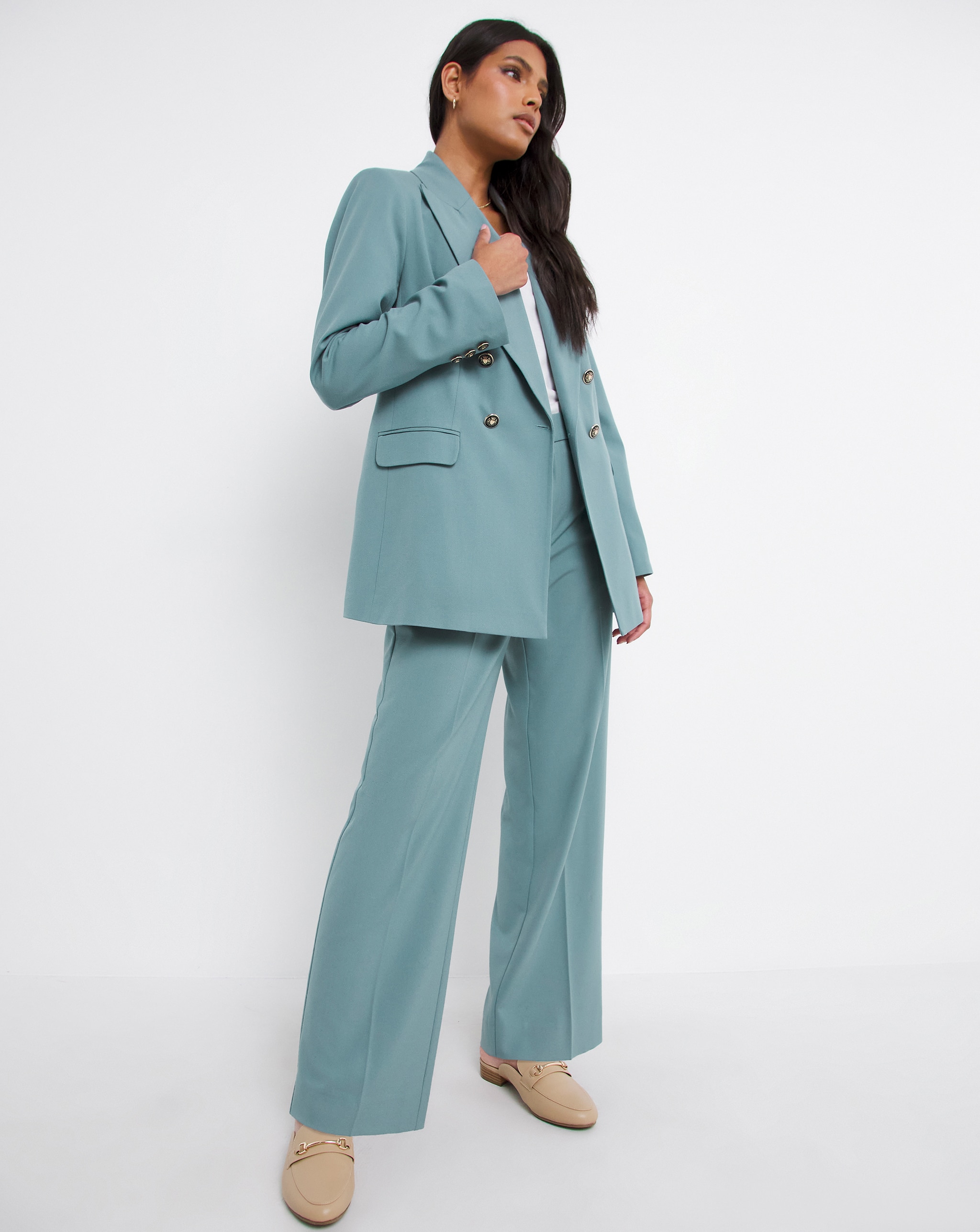 A person standing against a plain white background wearing a light teal tailored suit with a double‑breasted blazer and wide‑leg trousers.