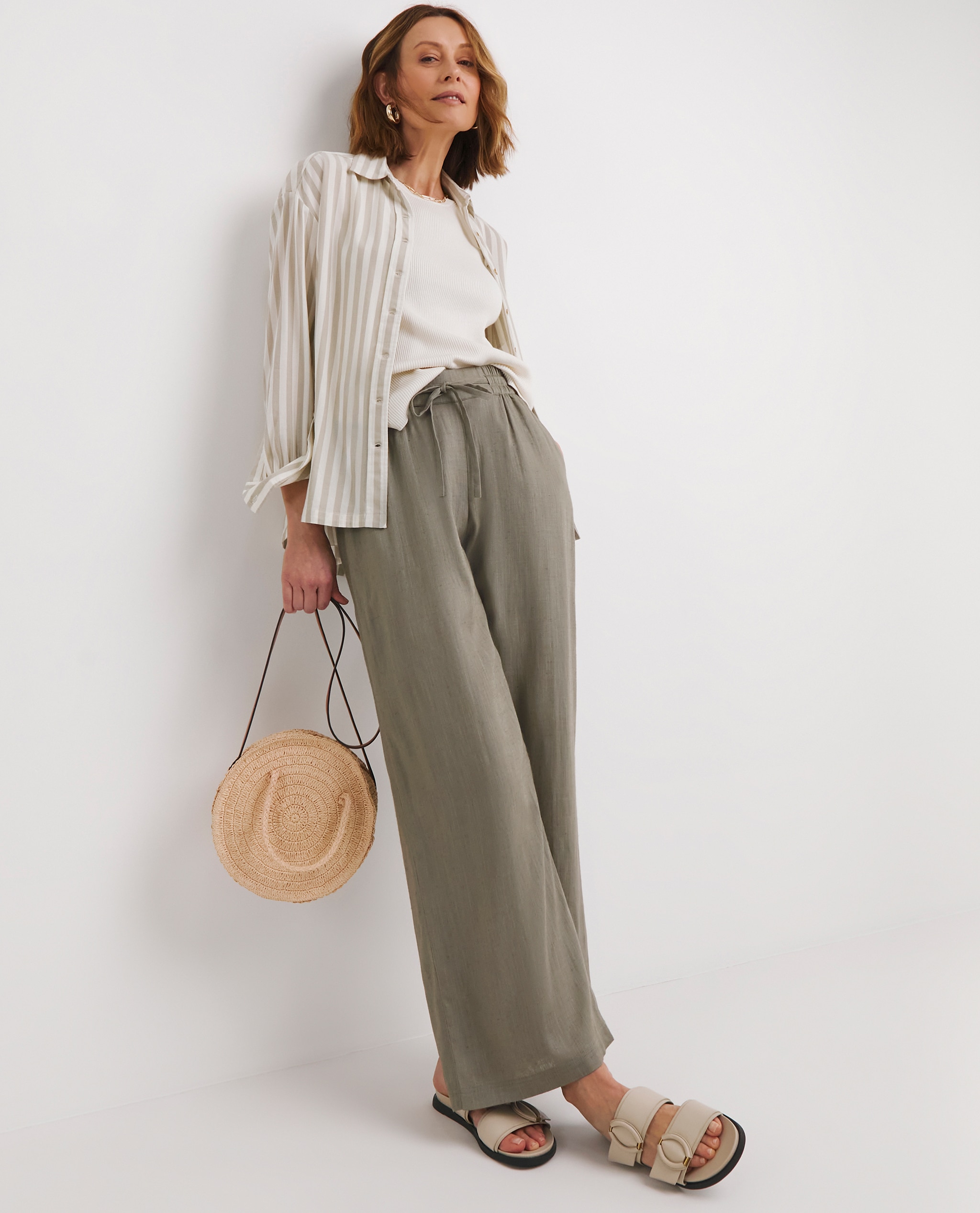 Model is wearing a pair of khaki linen mix wide leg trousers with a cream t-shirt and a stripe shirt over it with beige sandals.