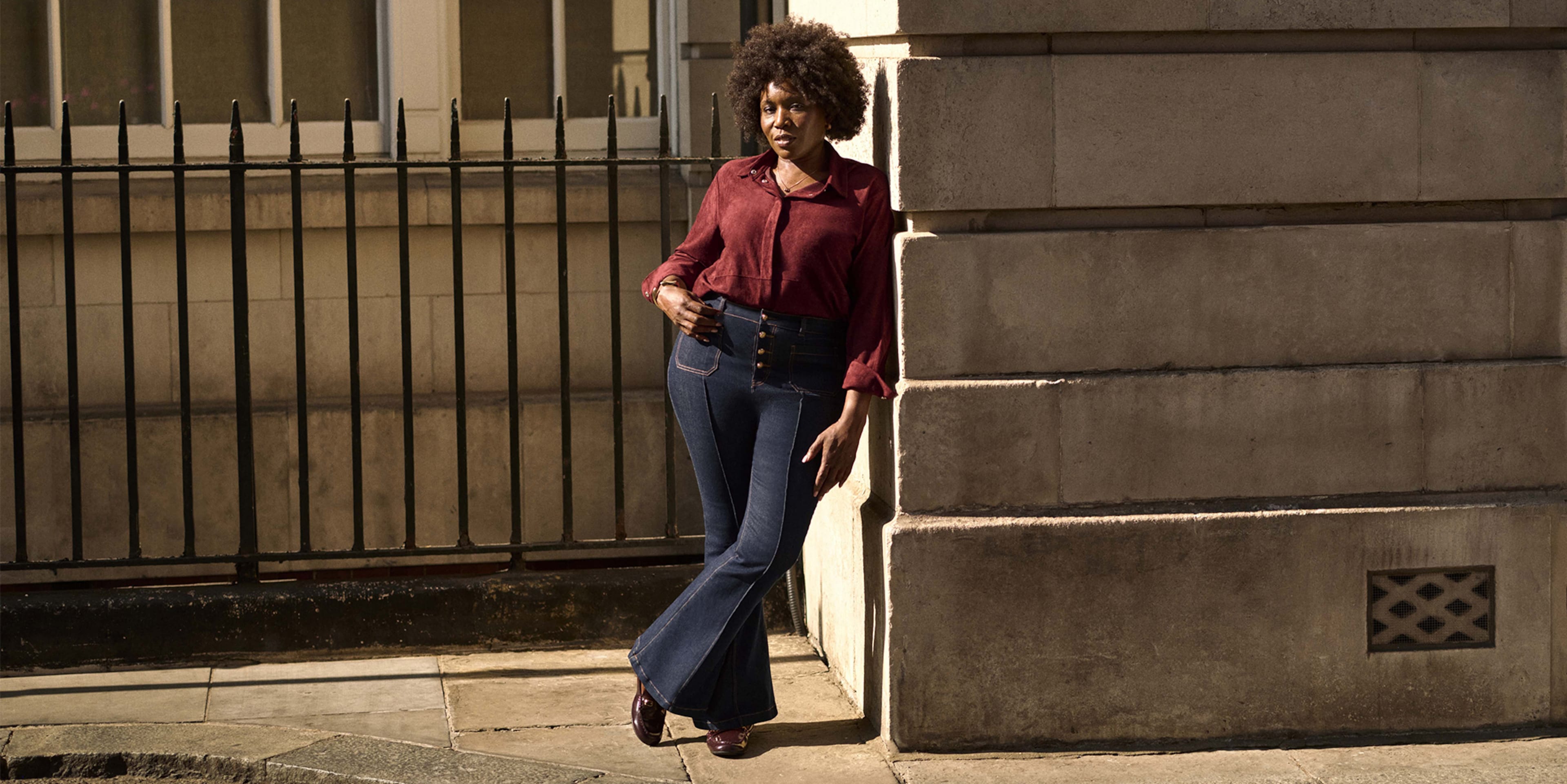 A lady stood outside leaning against a wall wearing a burgundy blouse with dark blue jeans.