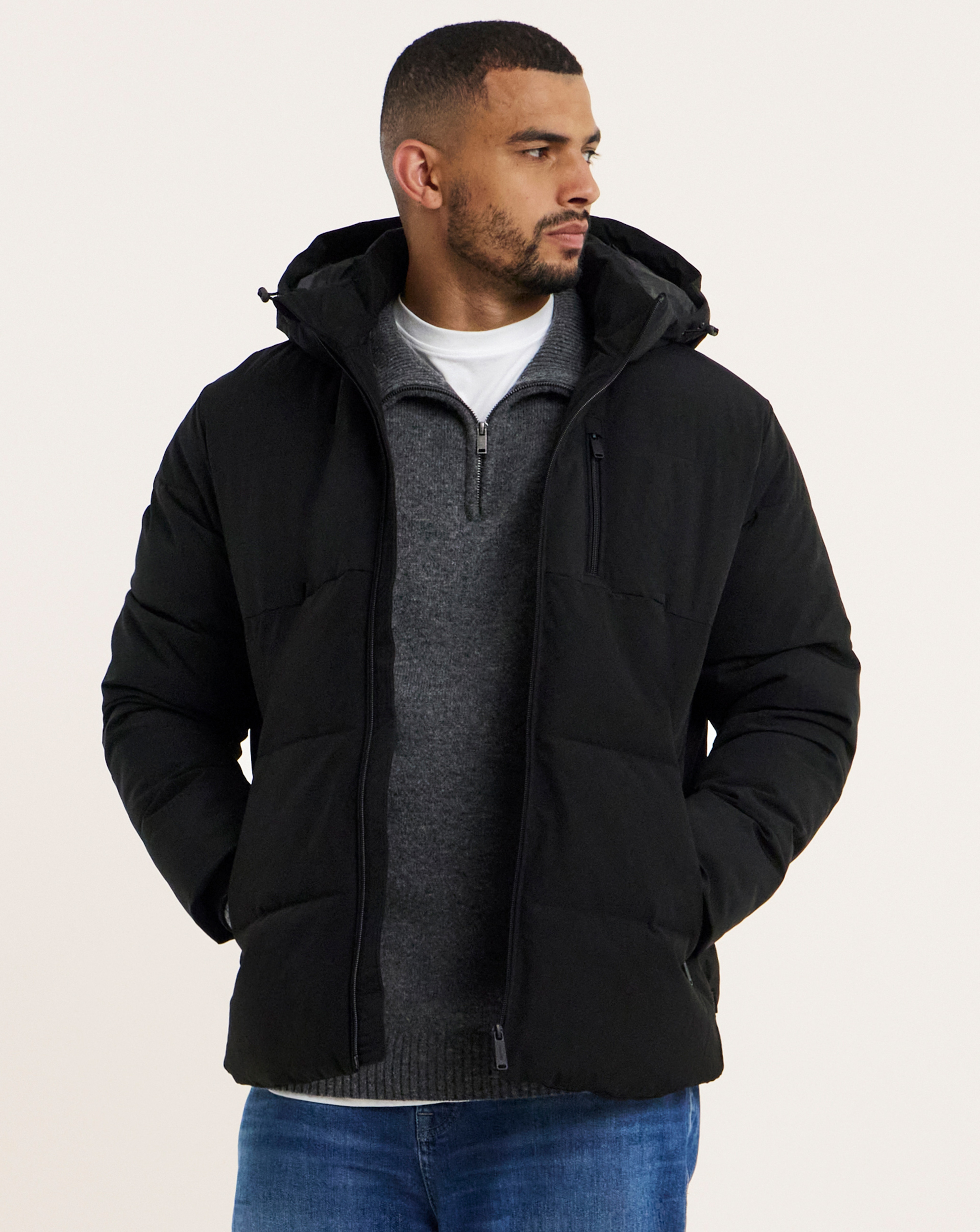 A man stood looking to the right wearing jeans, a white t-shirt, a grey zip up top and a black quilted coat.