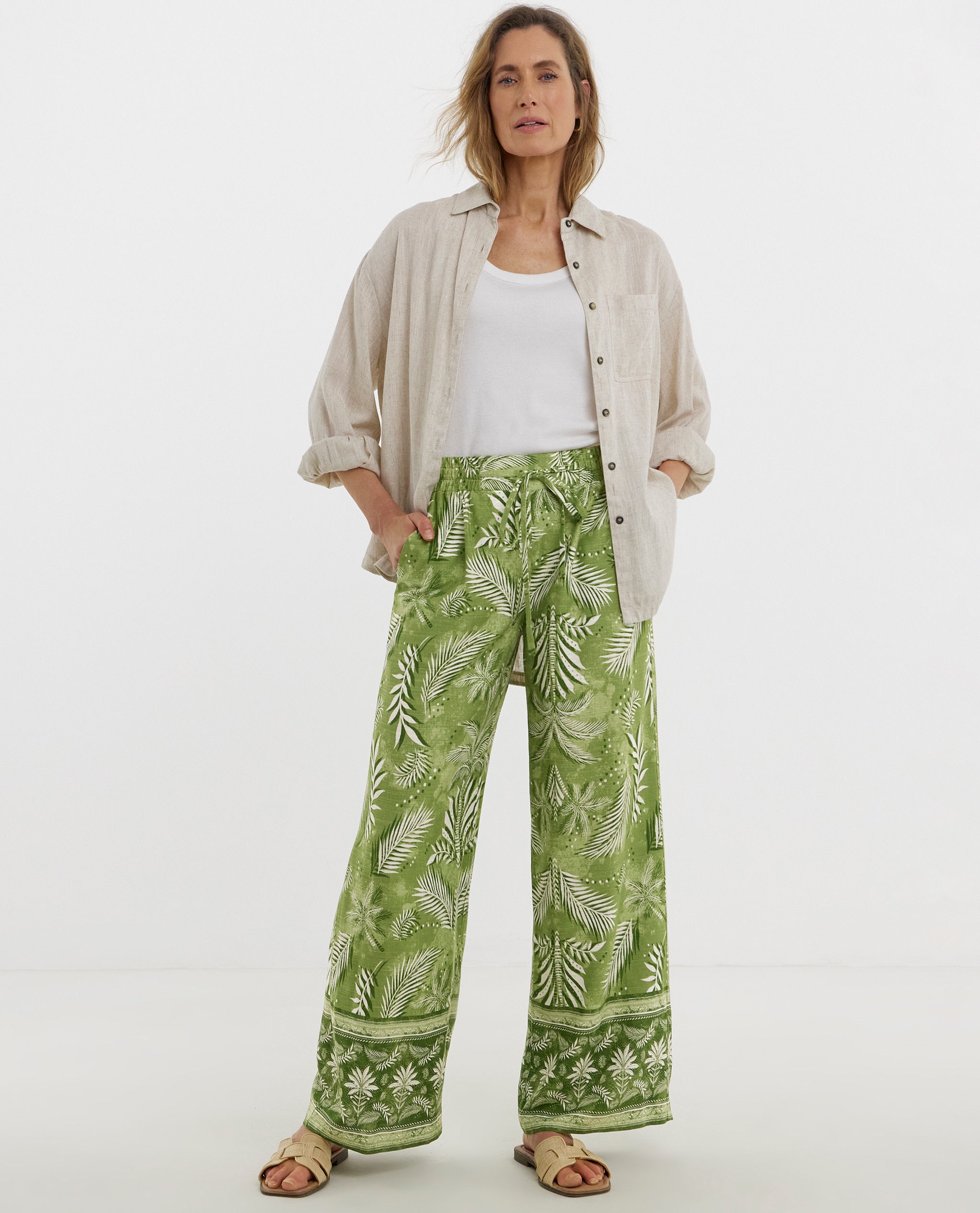 Model is wearing a white t-shirt with a beige shirt over it with a pair of green print linen mix wide leg trousers.