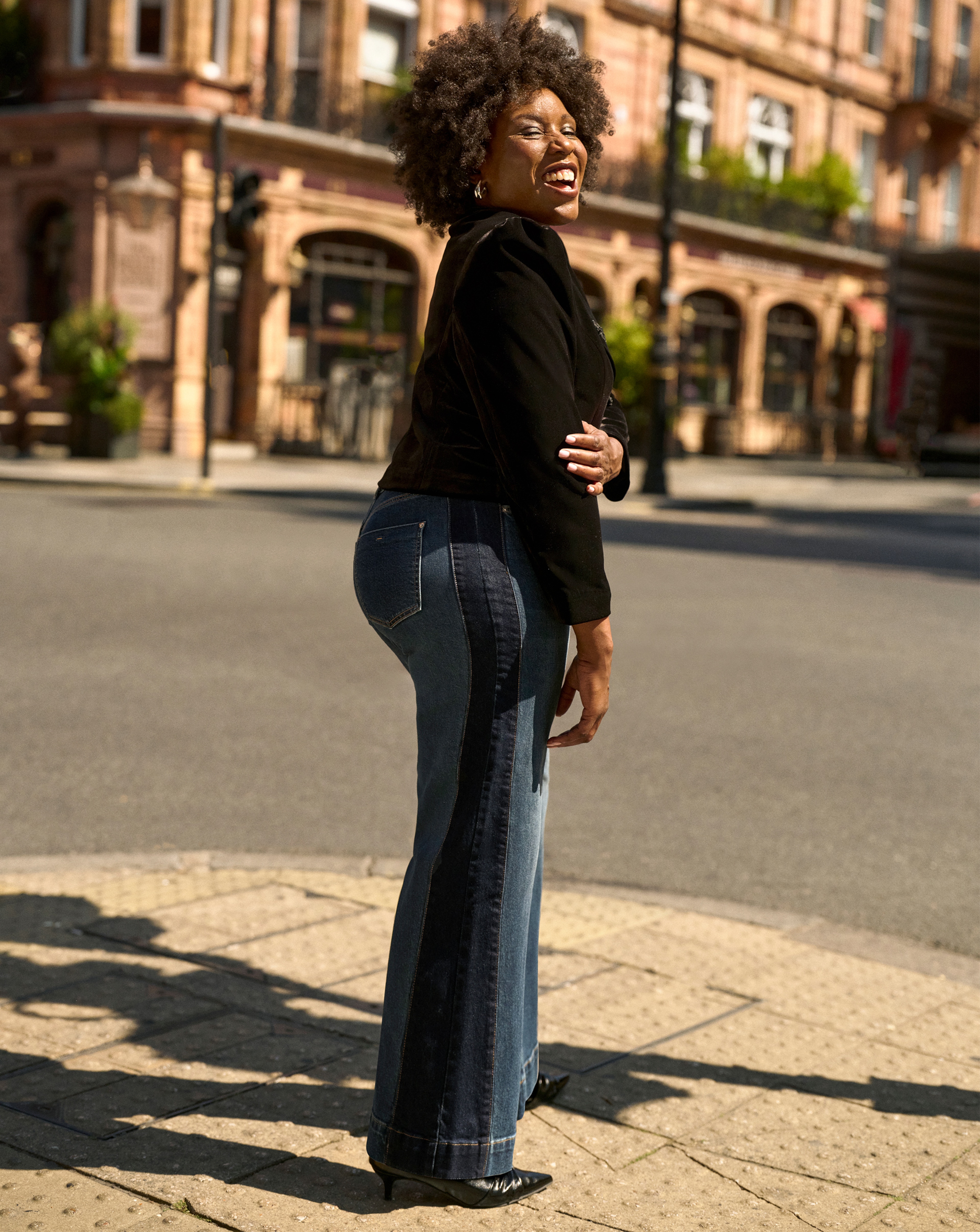 A woman stood on a corner of a street wearing a black jacket and jeans