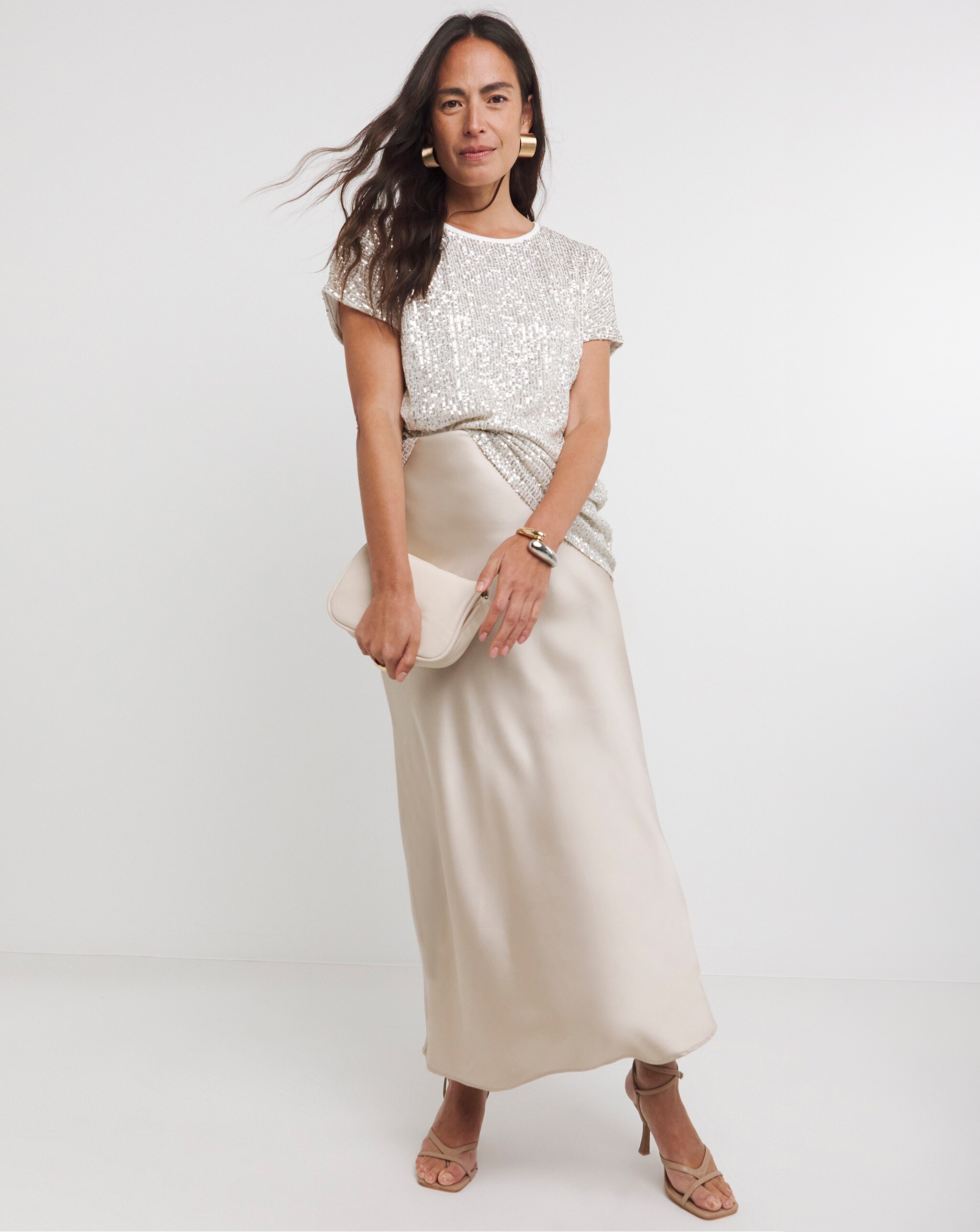 Person wearing a champagne sequin top with a satin maxi skirt, accessorised with a matching clutch and heeled sandals.