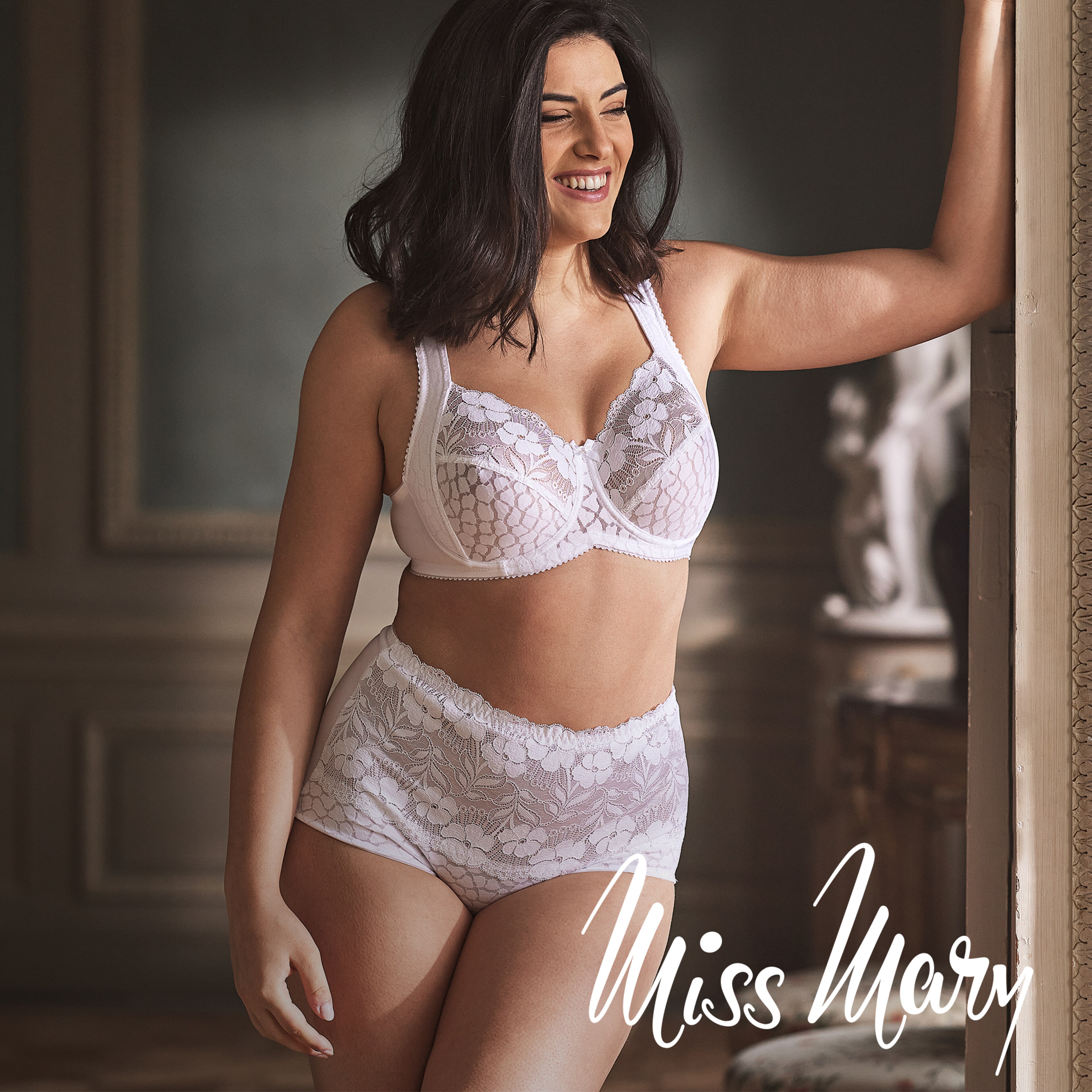 Model wearing a white Miss Mary of Sweden lingerie set, standing by a window.