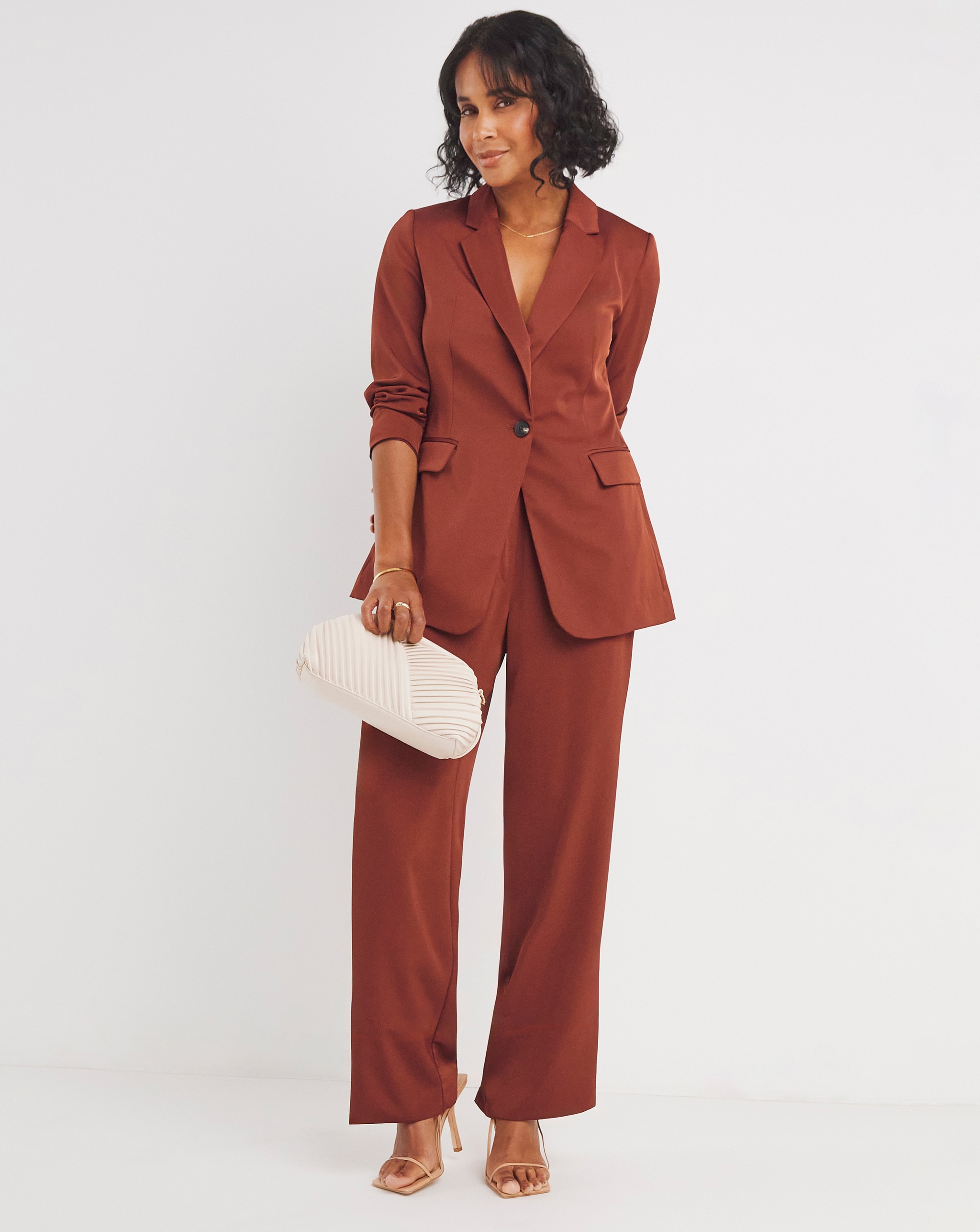 Person wearing a rust coloured tailored suit with a single button blazer and wide leg trousers, holding a cream clutch bag