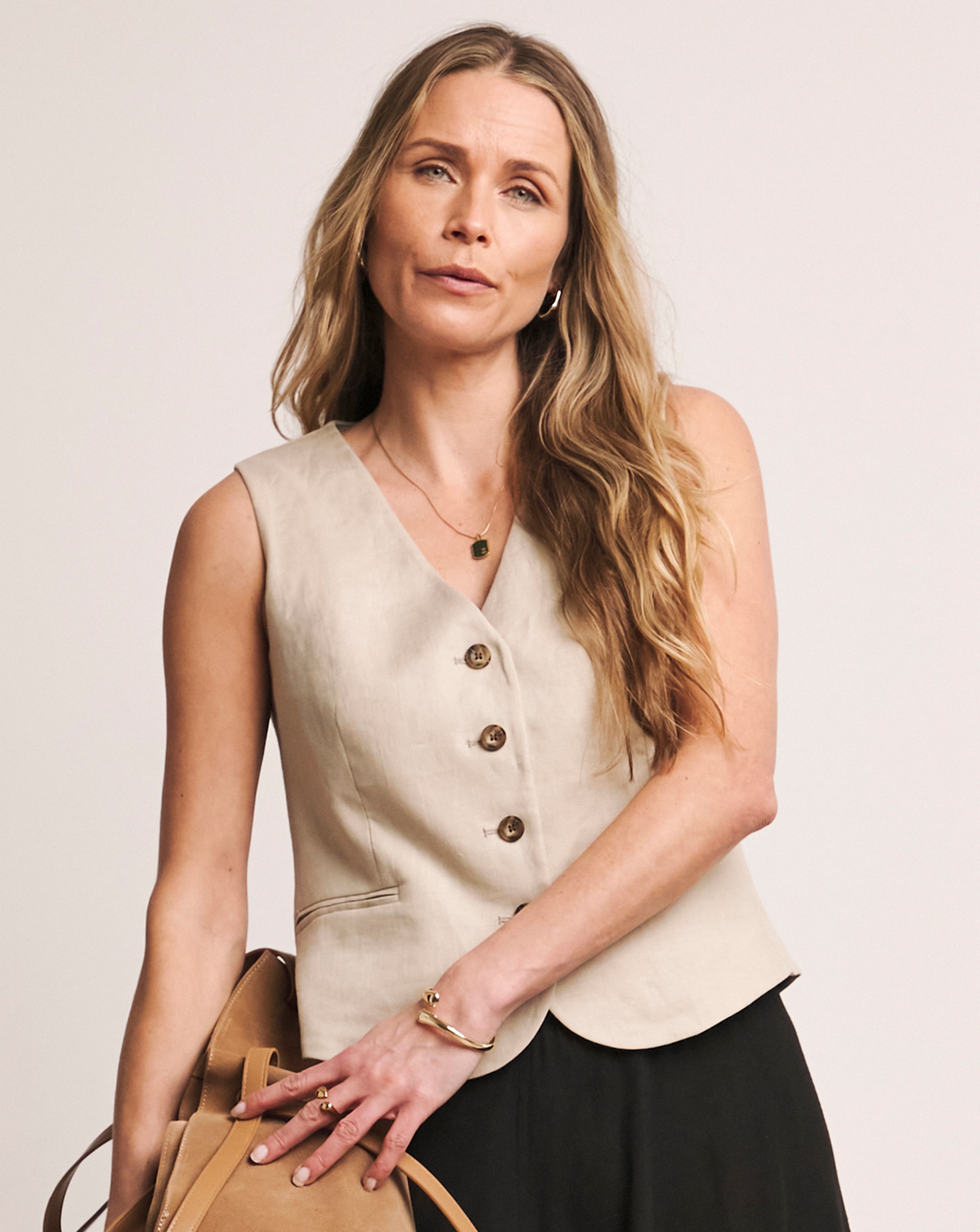 Person wearing a beige sleeveless vest, long black skirt, and holding a tan bag.