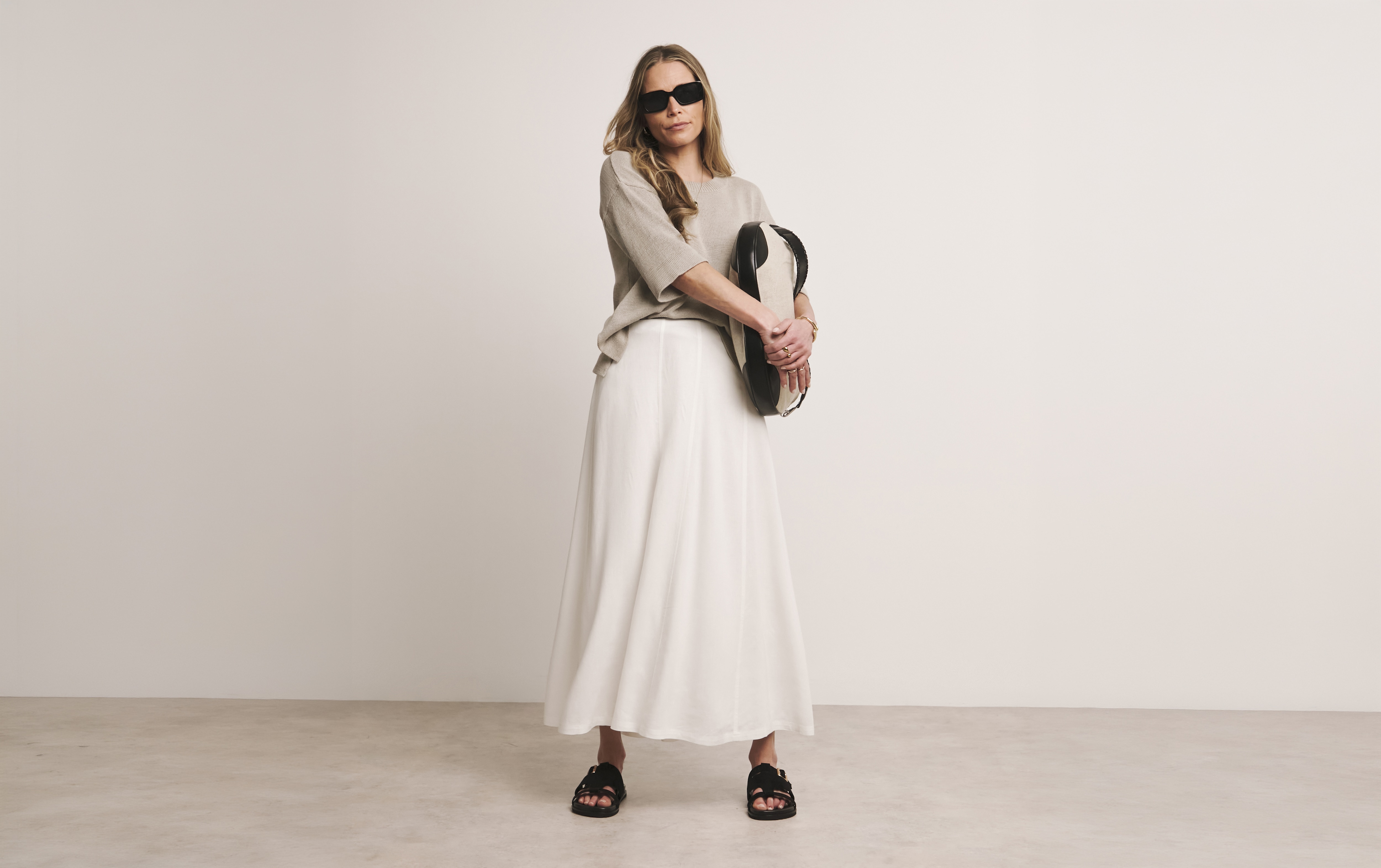 A lady in a beige room wearing a stone coloured top and white maxi skirt holding a bag.