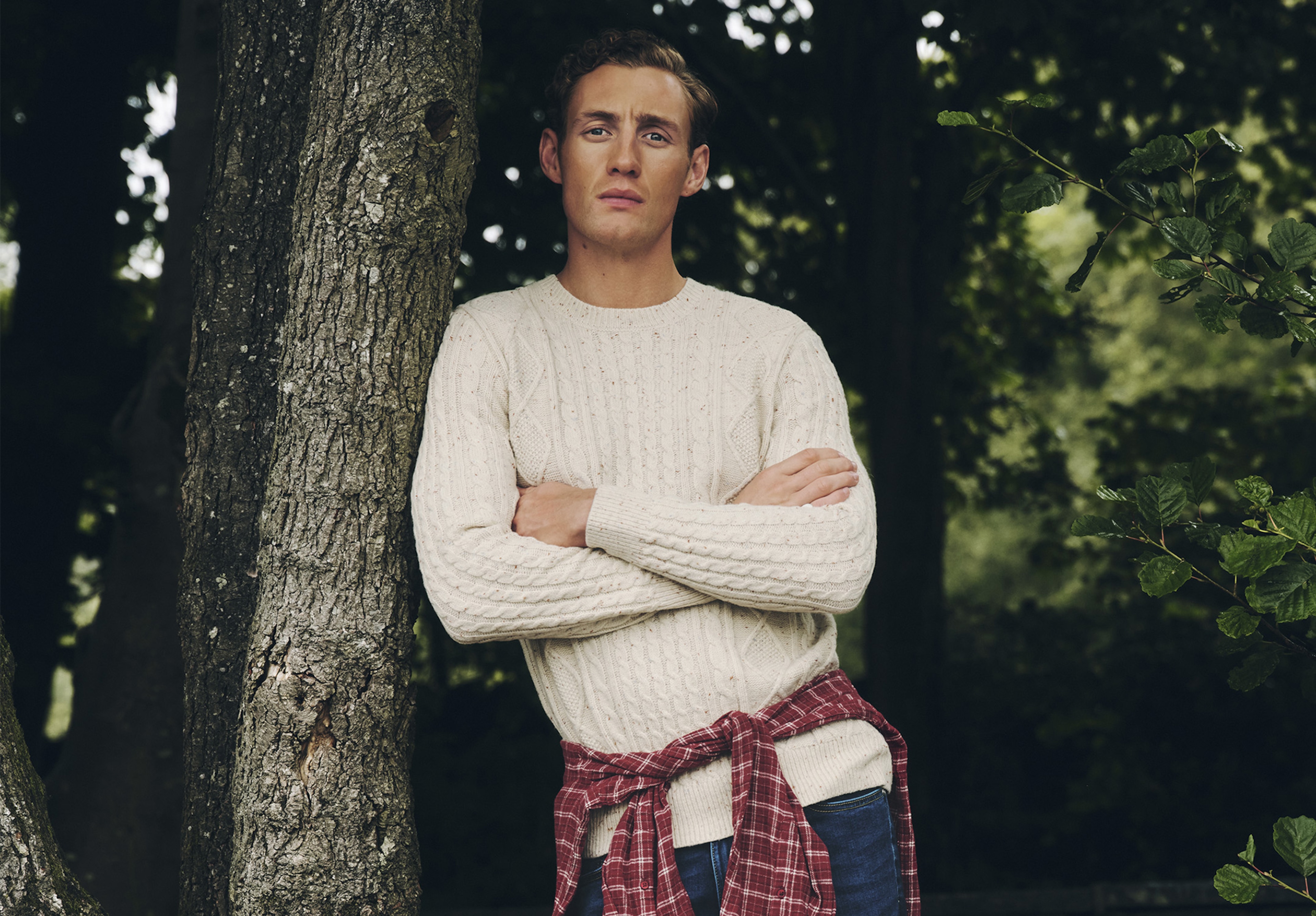 A man leaning against a tree wearing a cream knitted jumper with a red shirt tied round his waist.