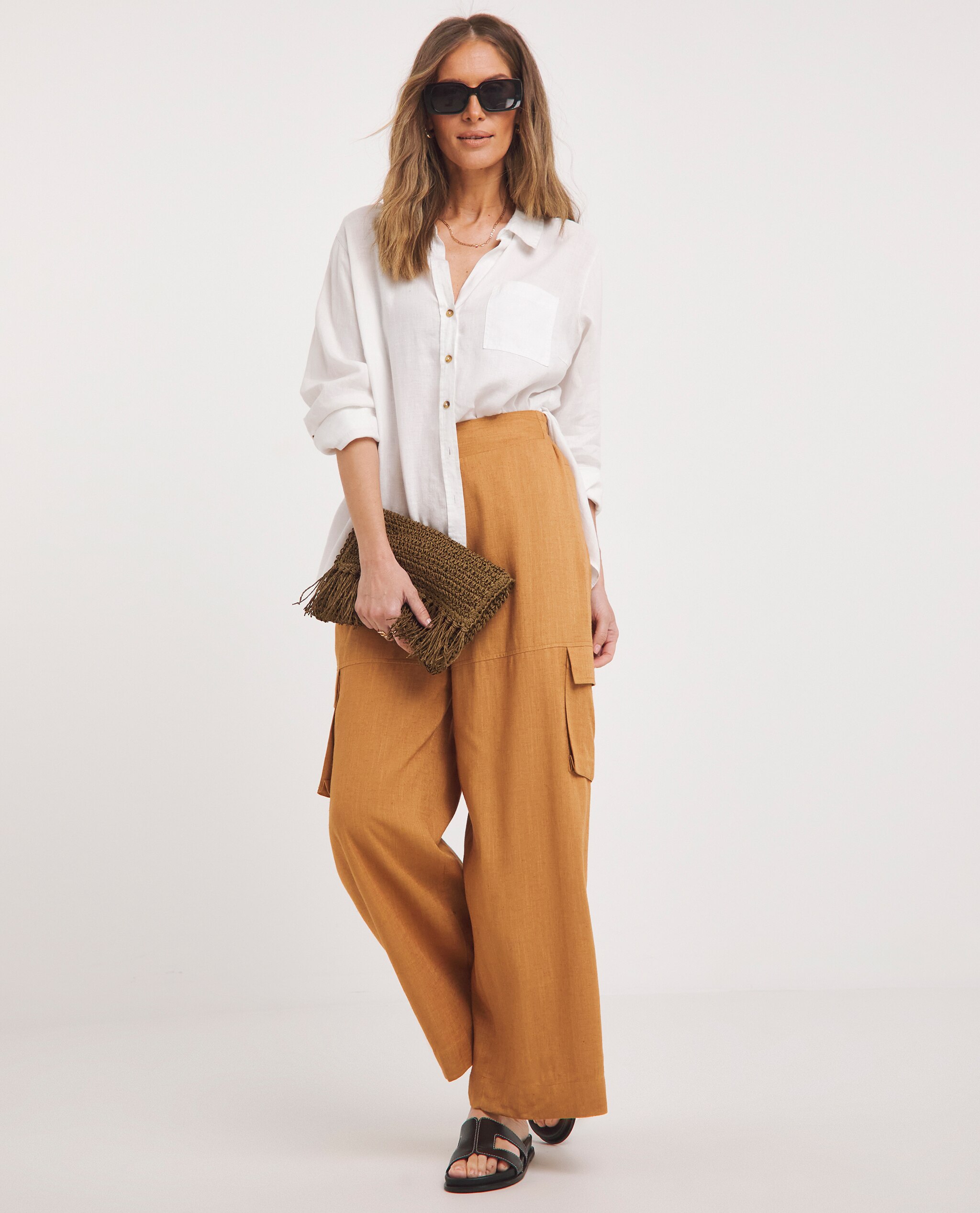 Model is wearing a white shirt with a pair of burnt orange linen trousers with a brown crochet bag and black sandals.