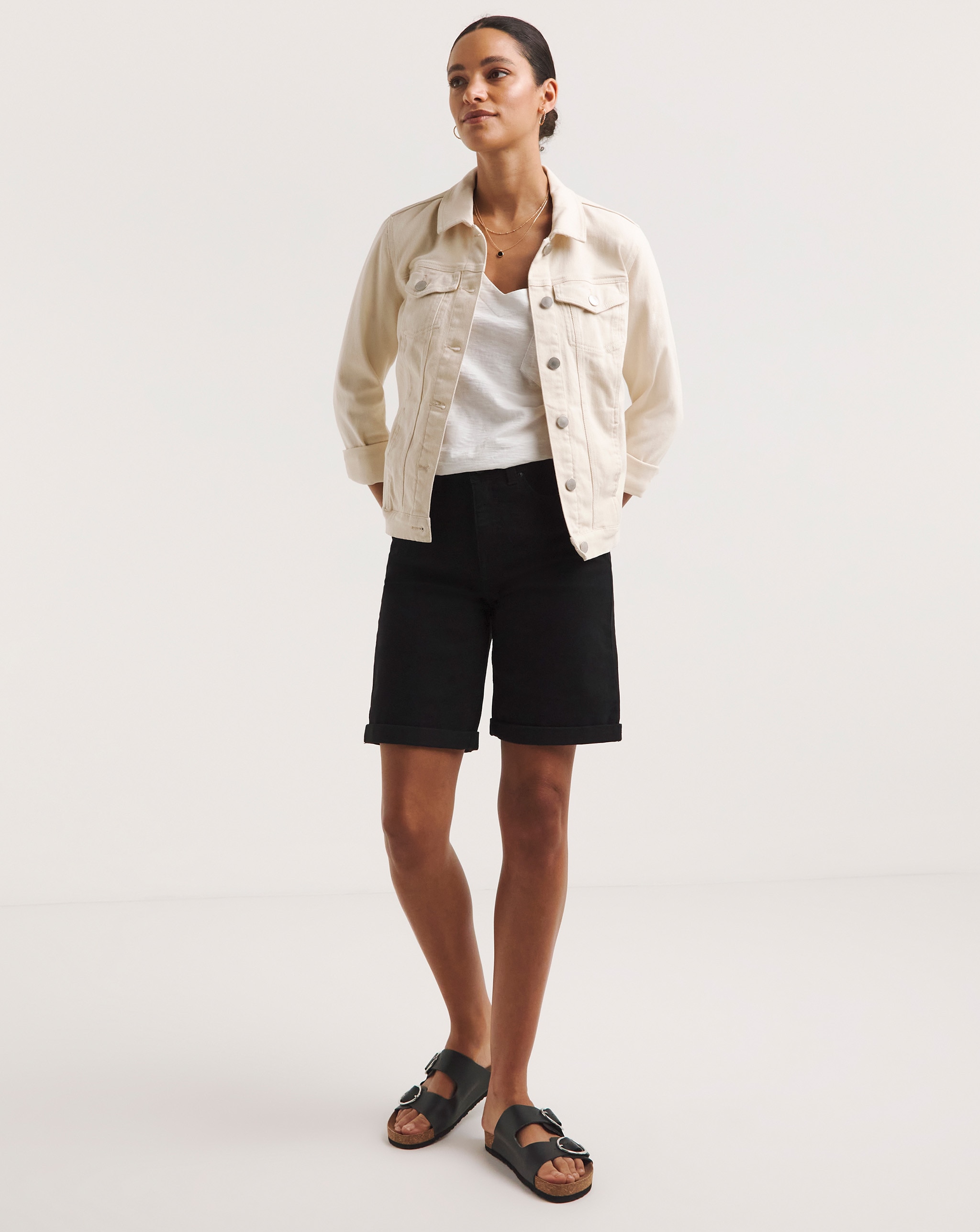 Person wearing a cream denim jacket over a white top, styled with black knee-length shorts and black buckle sandals.