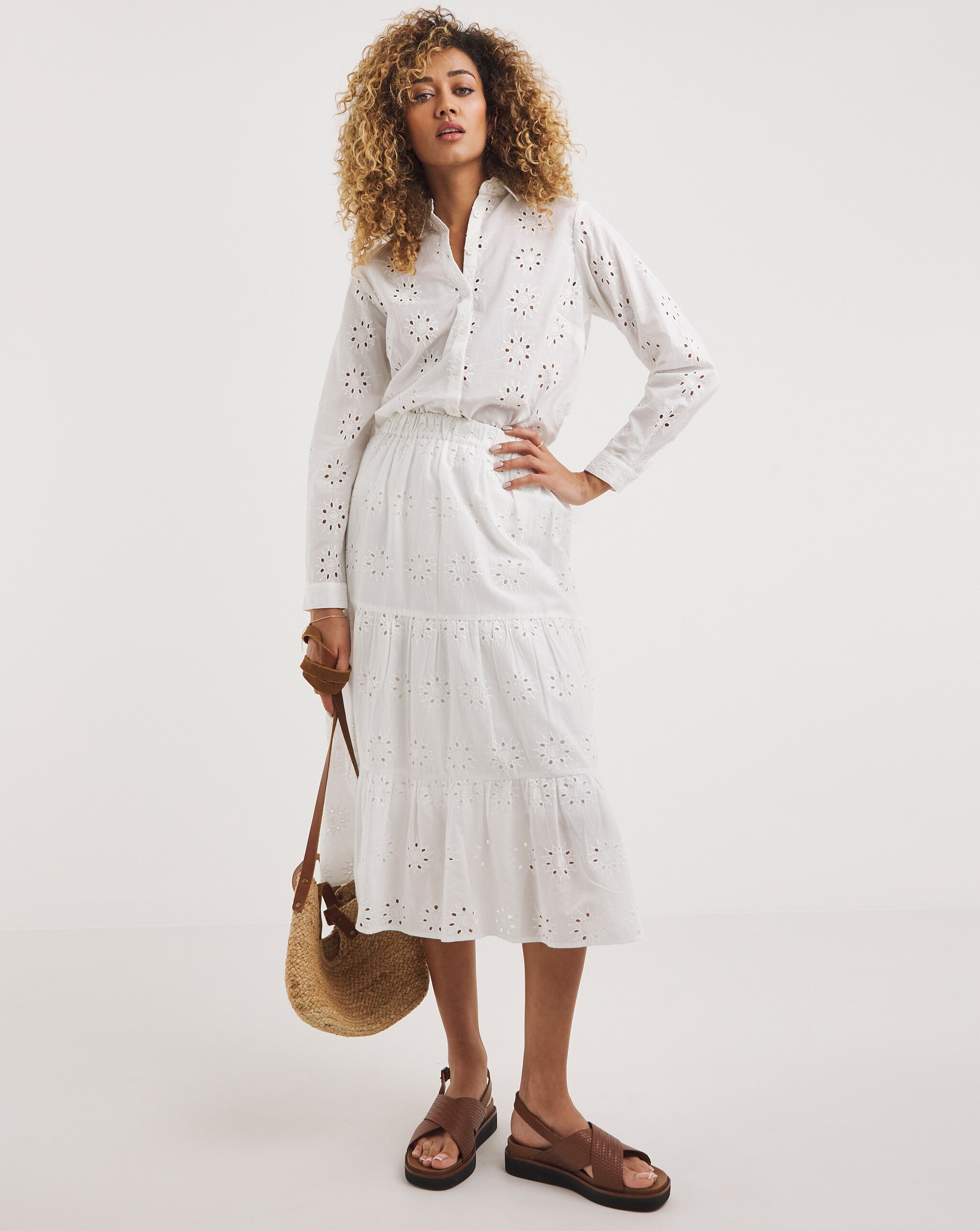 Person wearing a white broderie anglaise shirt and matching tiered midi skirt with brown sandals and a woven shoulder bag.