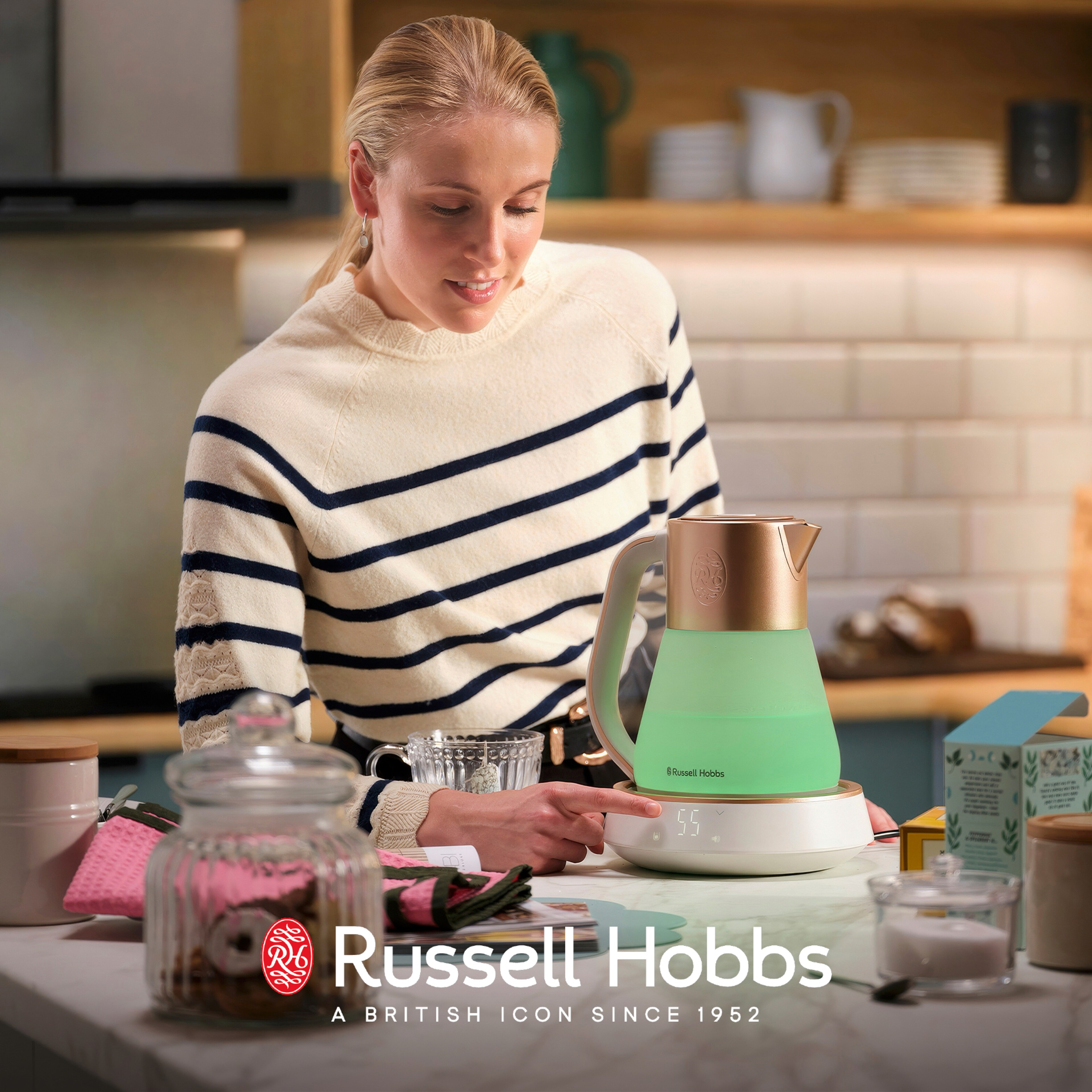 Person in a striped sweater using a Russell Hobbs Calm kettle on a kitchen countertop with cups and jars nearby.