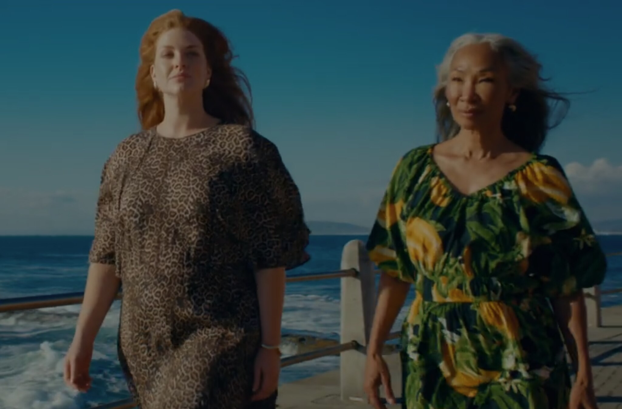 Two ladies walking next to the sea wearing dresses and the wind is blowing through their hair.