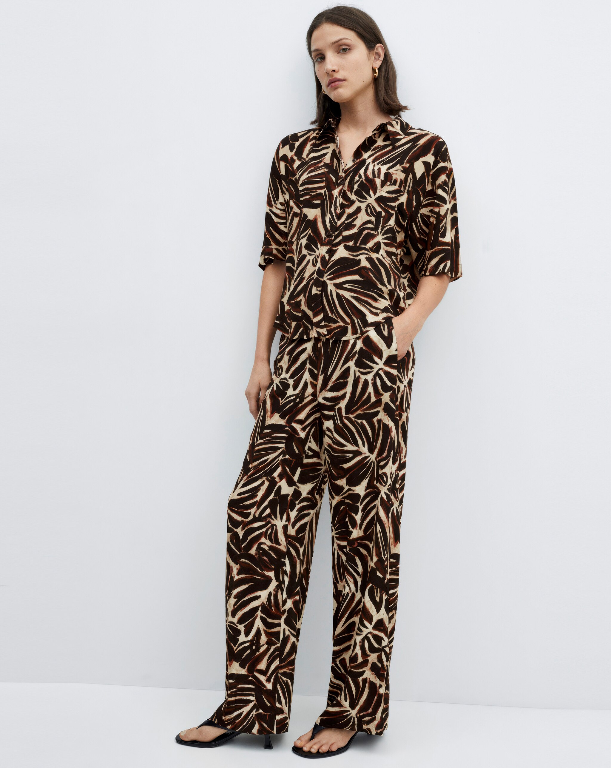 Person standing in a brown and cream patterned short‑sleeve shirt and wide‑leg trousers.