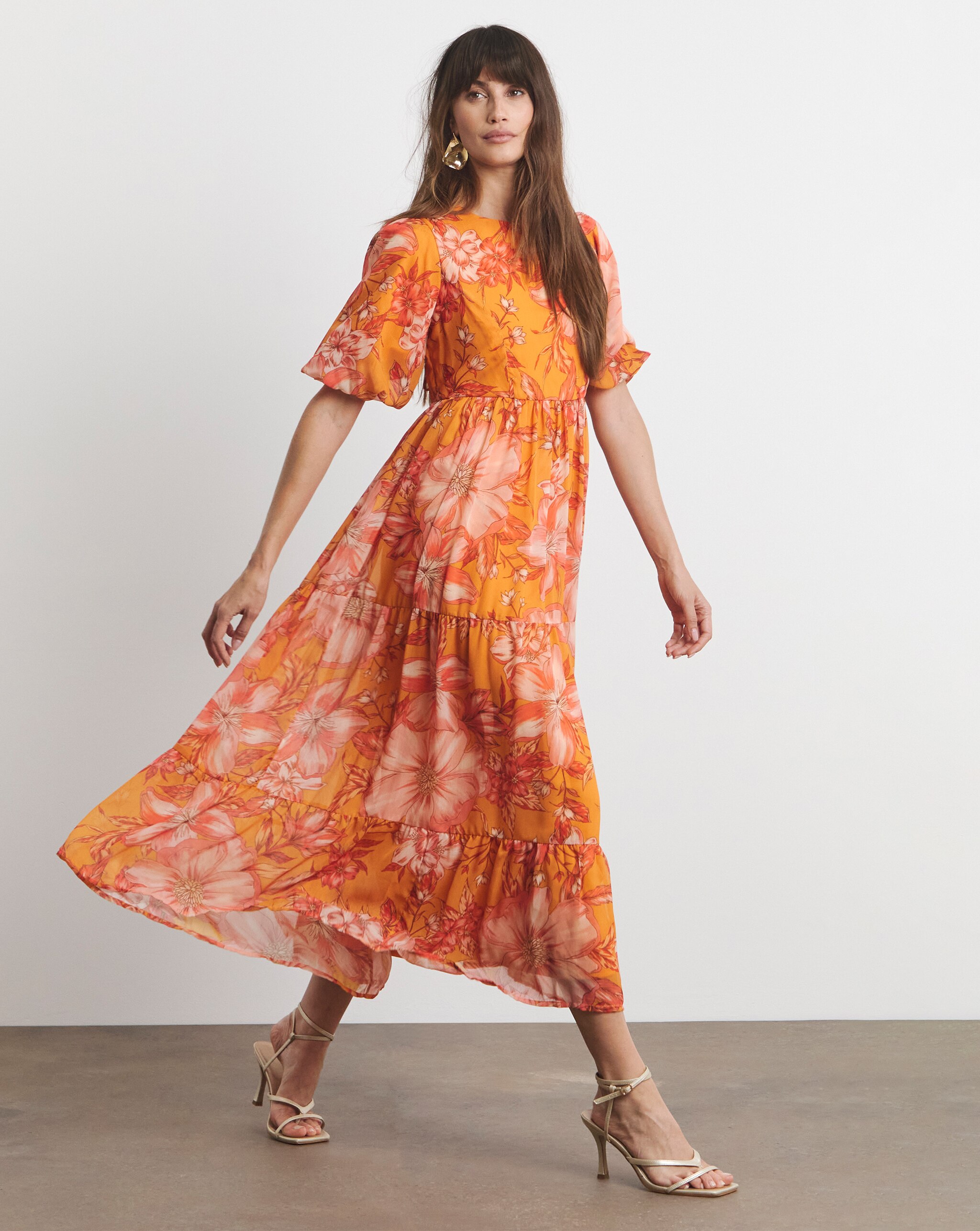 Person wearing a bright orange Joanna Hope maxi dress with short flutter sleeves and gold strappy heels in a studio setting.