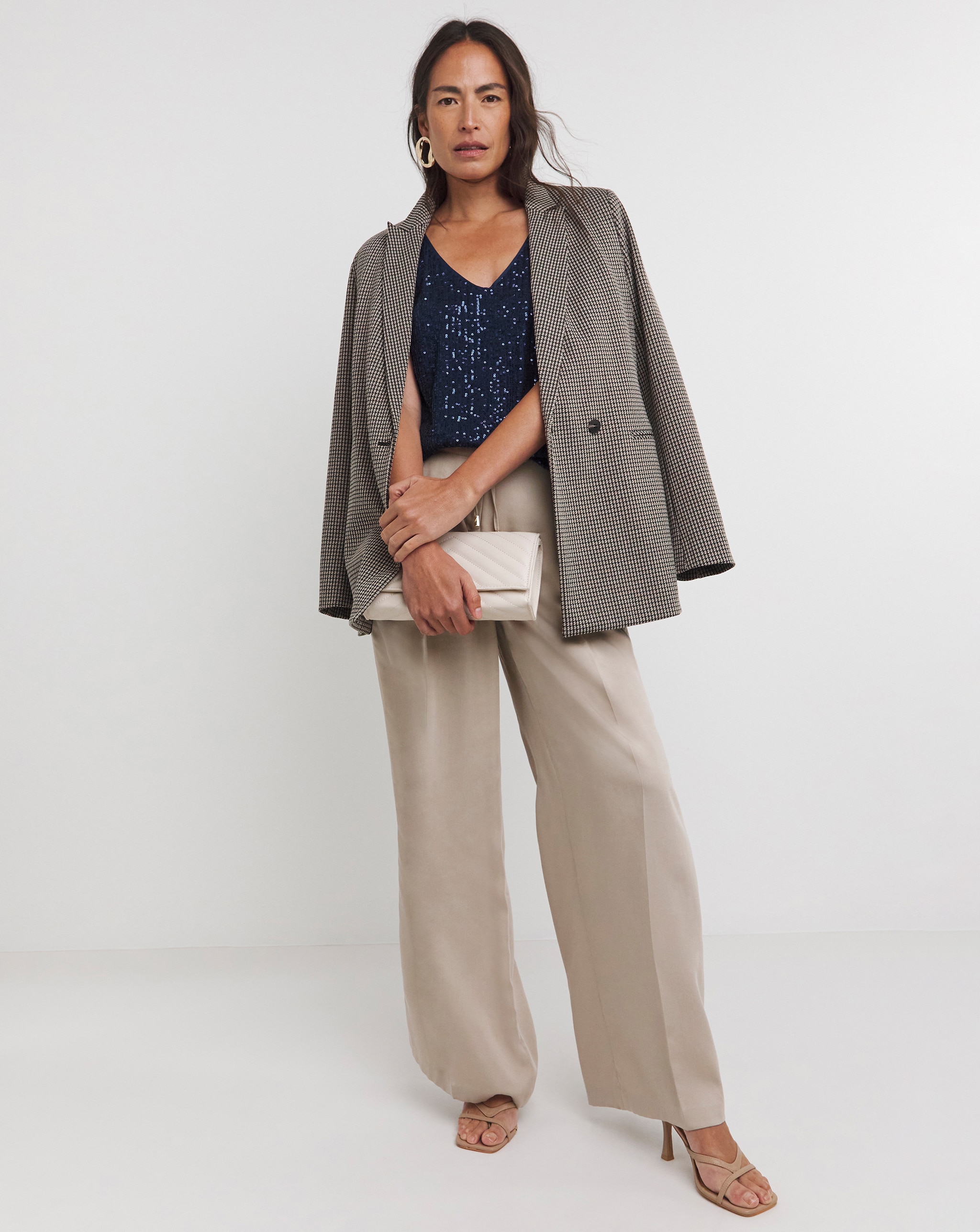 Person standing in a checked blazer draped over the shoulders, a navy sequin top, beige wide‑leg trousers, and holding a light clutch.