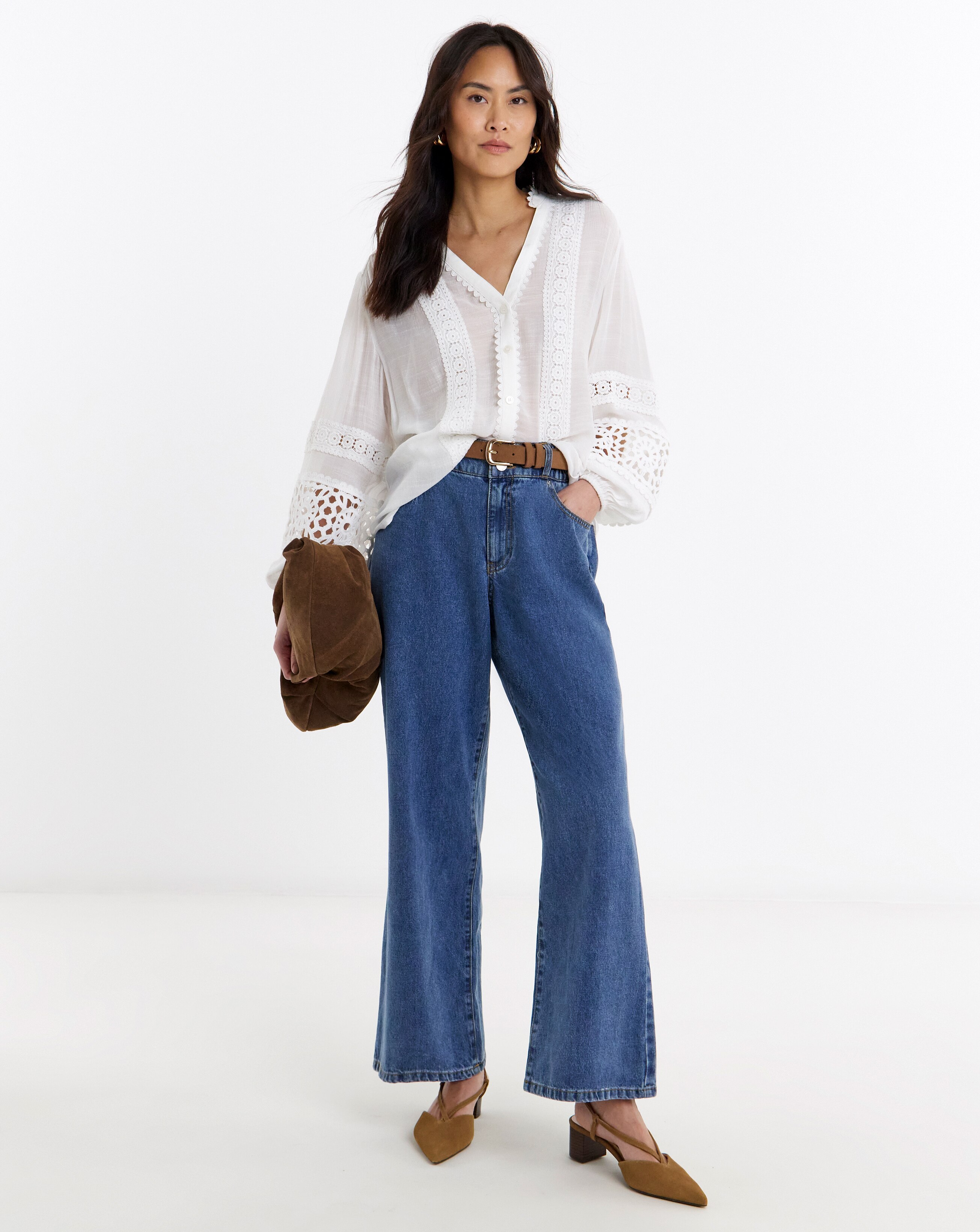 Person standing in a studio, wearing a white lace-trim blouse with wide-leg blue jeans, tan heels, and carrying a brown suede clutch.