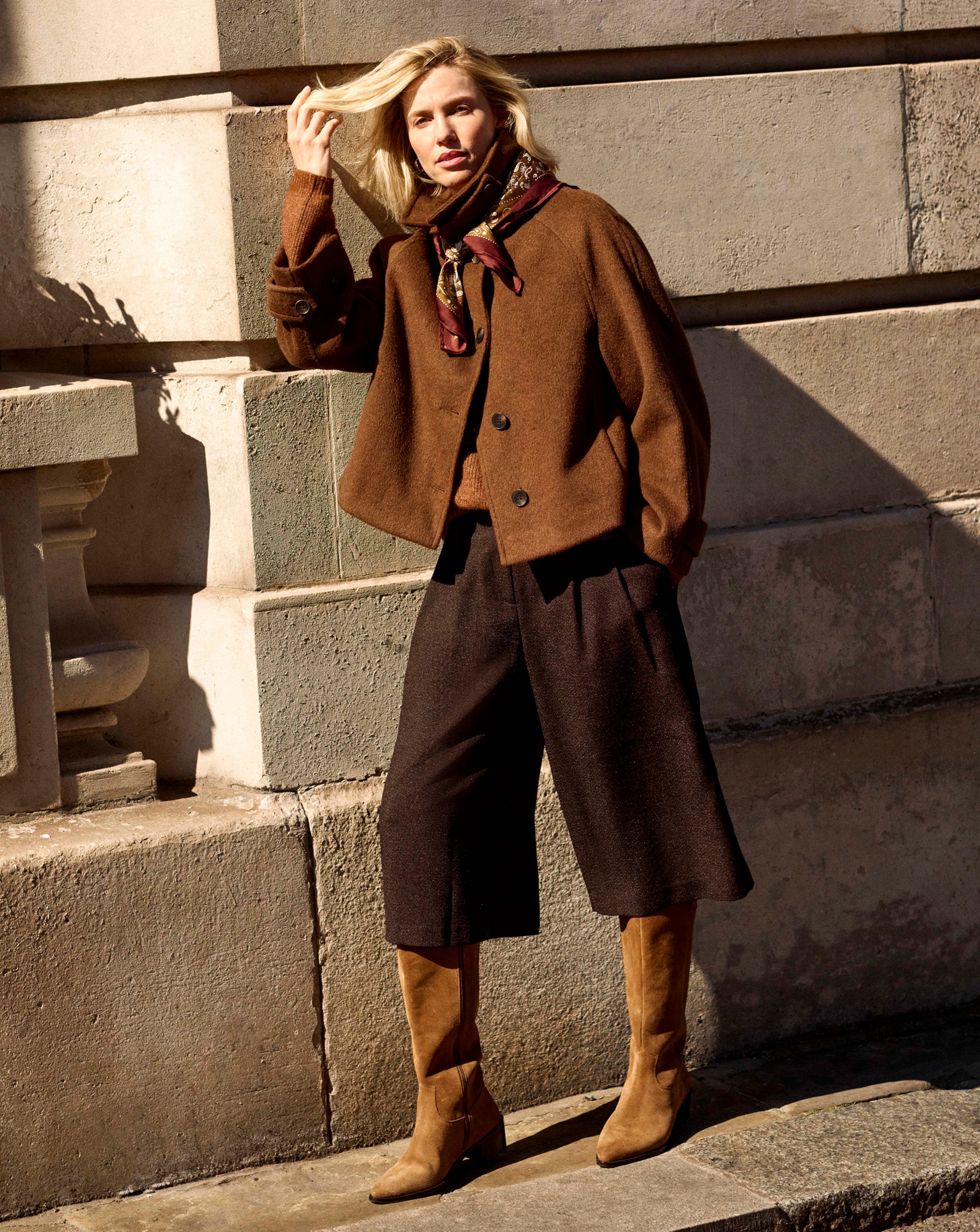 A woman leaning against a wall wearing a brown short funnel neck jacket, brown culottes and light brown boots