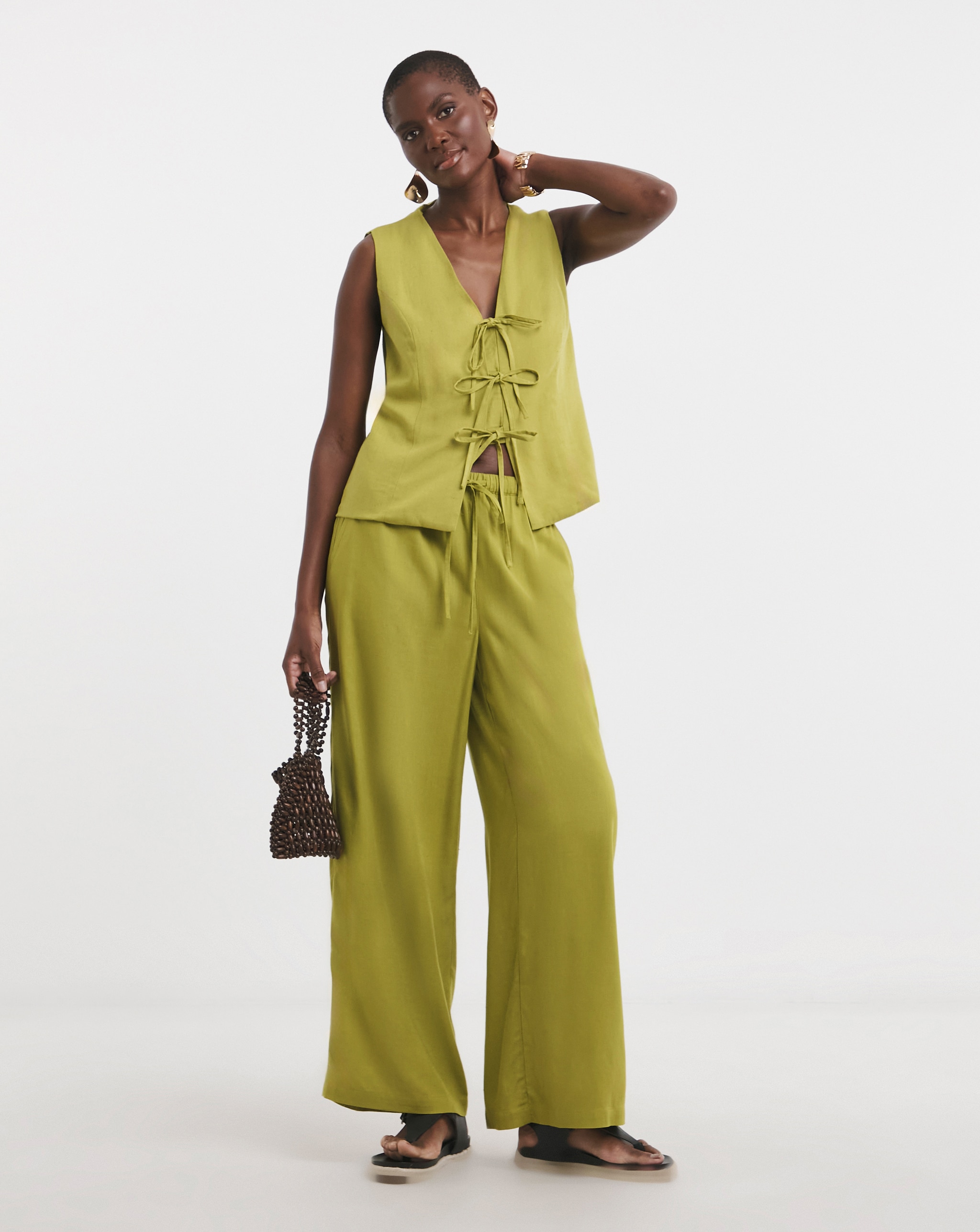 Person standing in a light green lace‑up top with matching wide‑leg trousers, holding a small beaded bag.