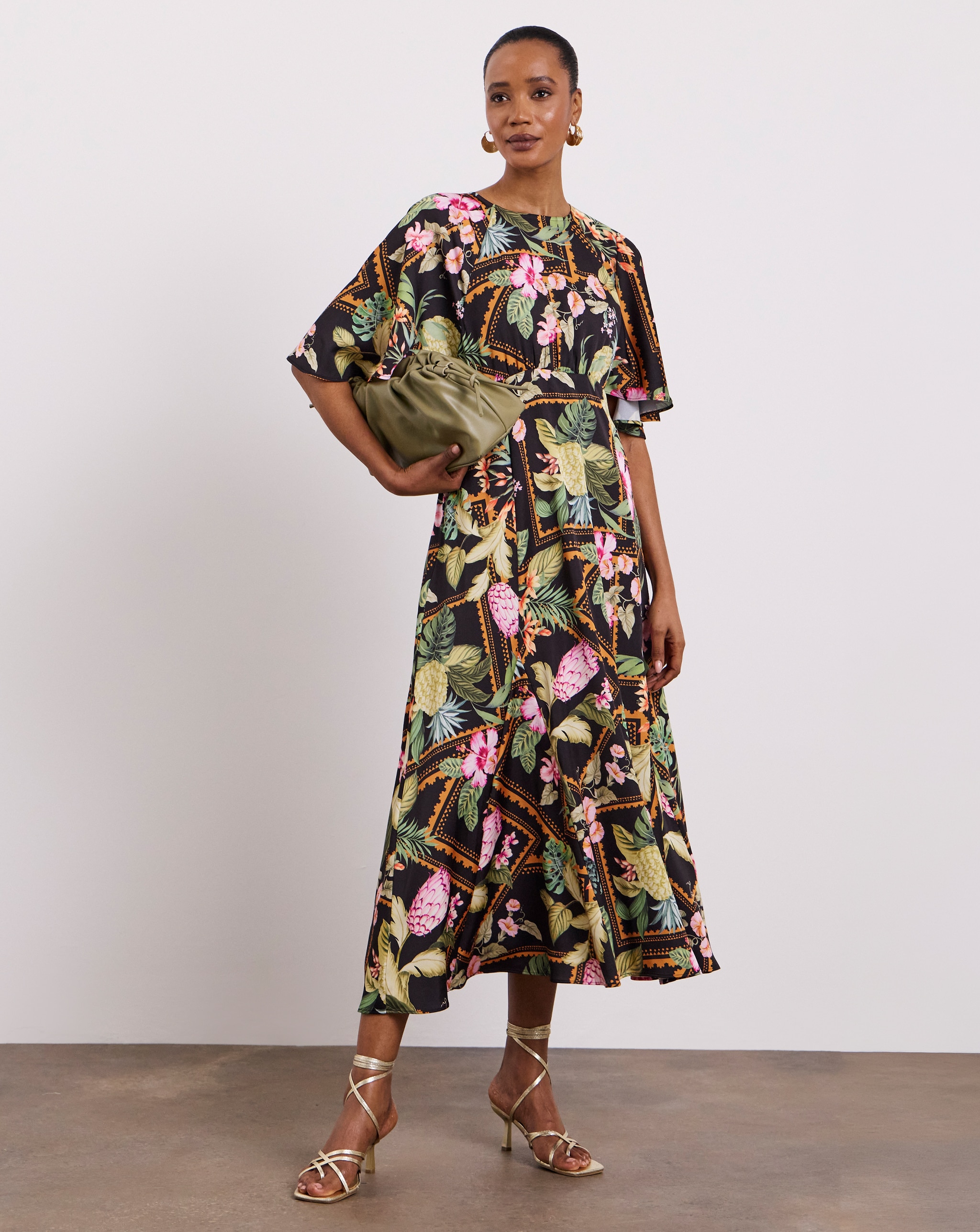 Person wearing a Joanna Hope tropical-print midi dress with flowing sleeves, gold strappy sandals, and an olive clutch bag.