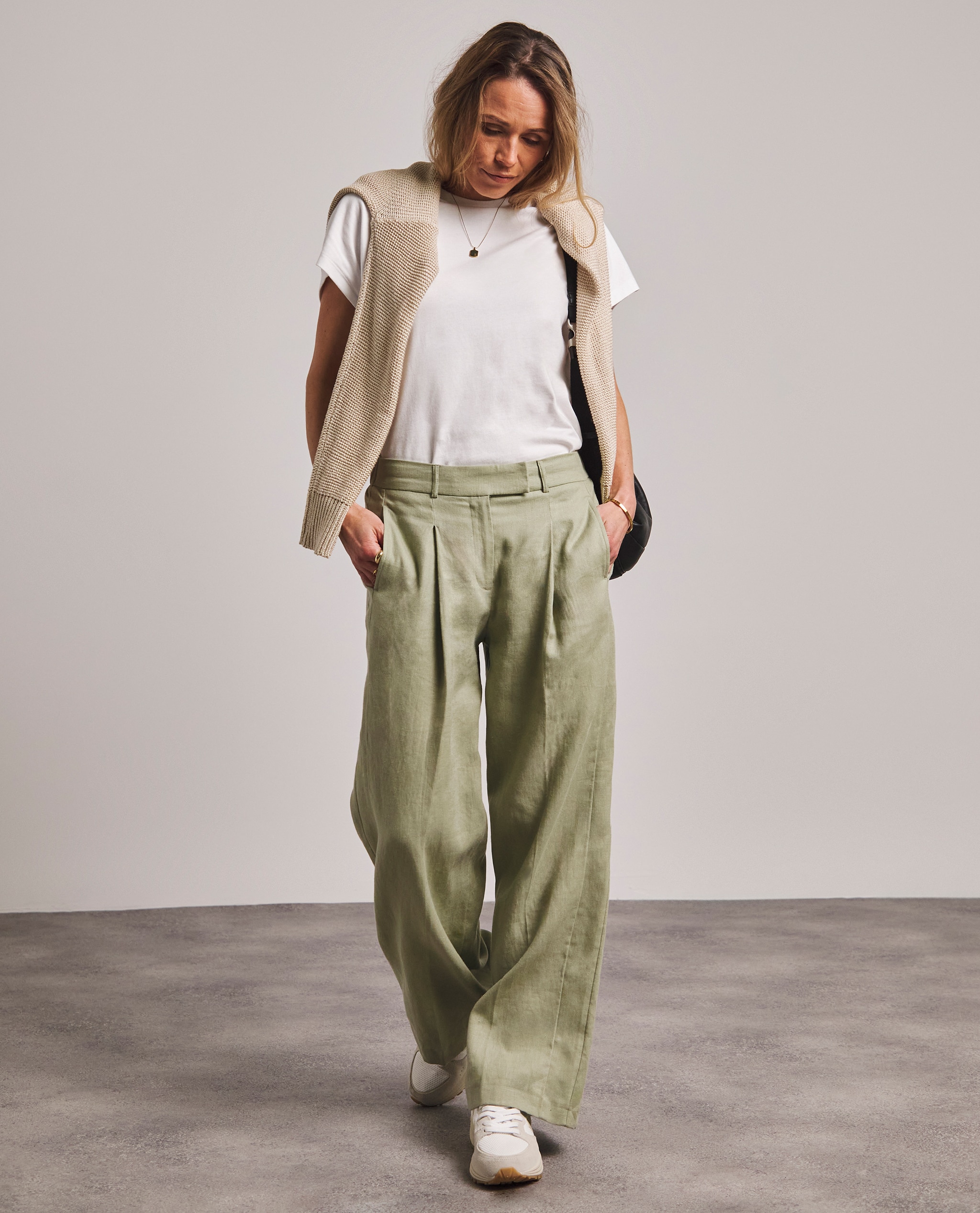 Model is wearing a white t-shirt with a cream jumper over her shoulders with a pair of green linen trousers.