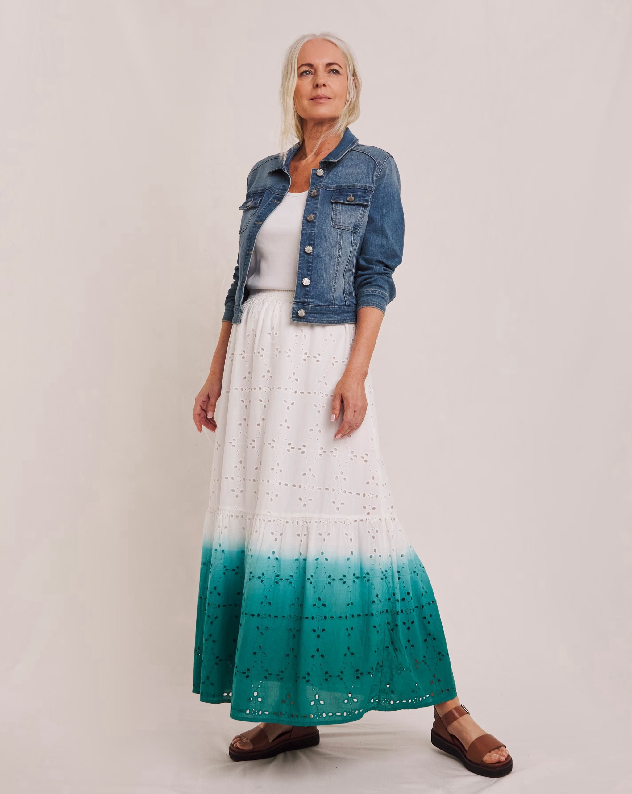 Person wearing a fitted blue denim jacket over a white top and white ombré maxi skirt with a teal hem, paired with brown sandals.