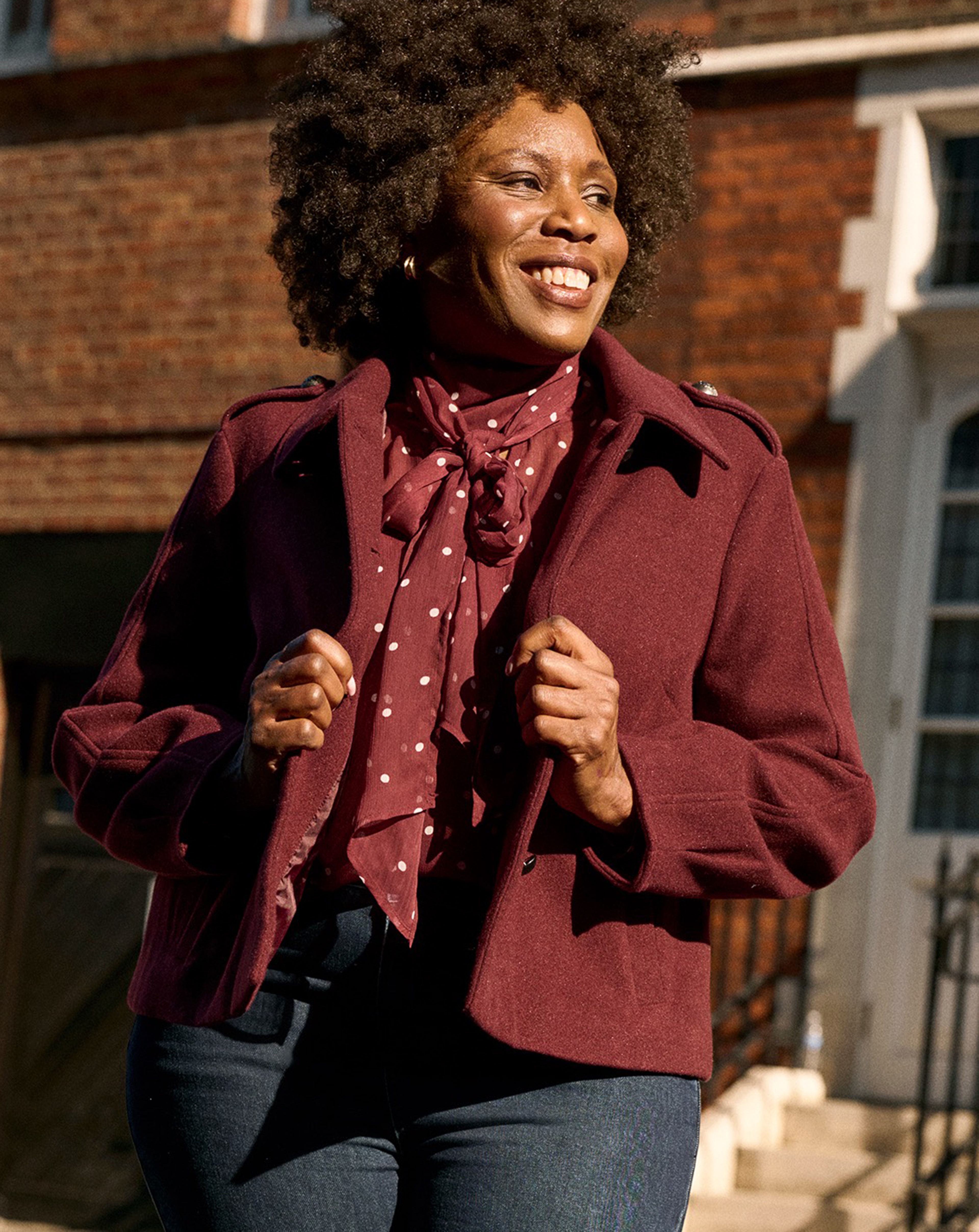 Person wearing a burgundy button front jacket and polka-dot neck scarf with dark jeans, standing outdoors in front of brick buildings.