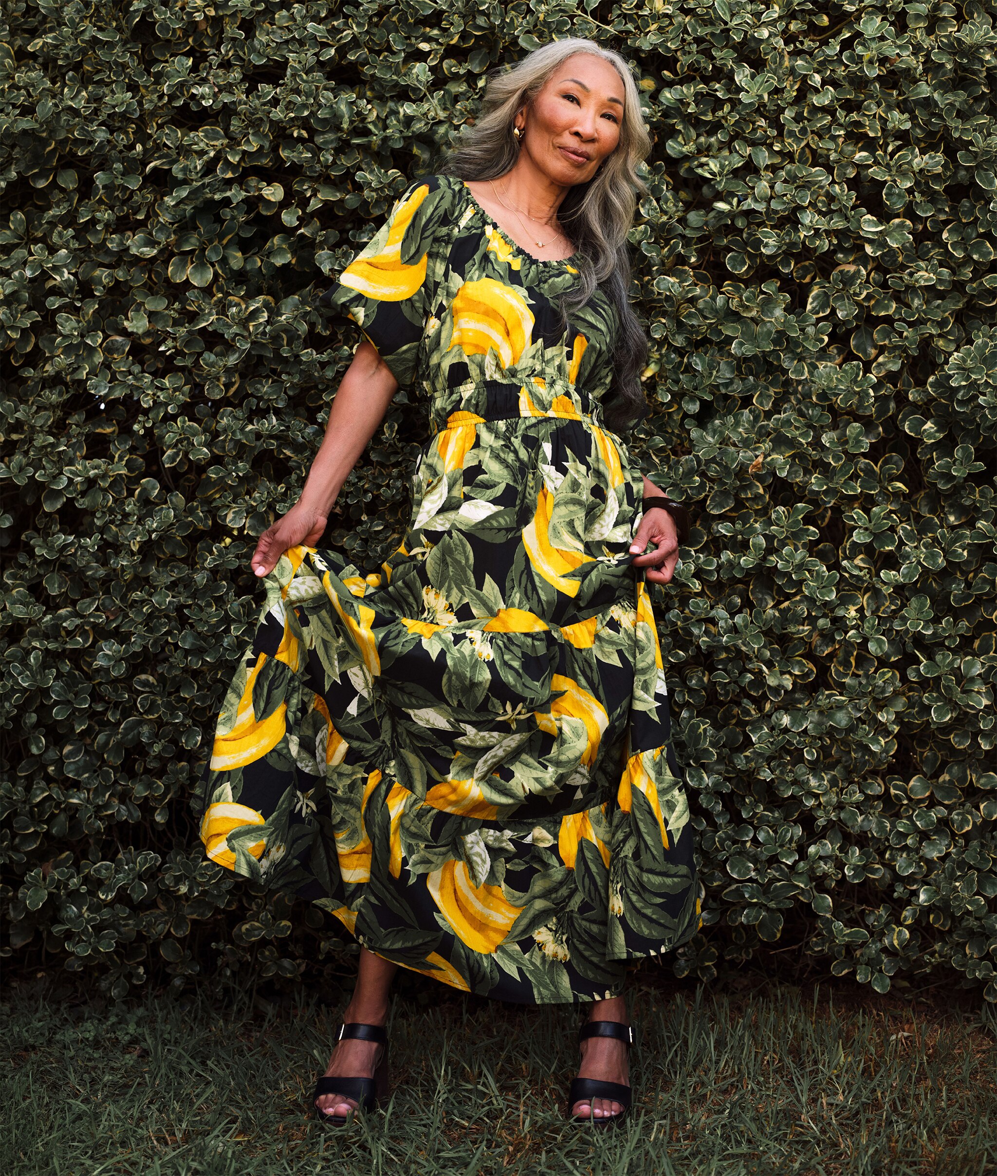 A lady stood in front of a hedge wearing a print dress with bananas on it.