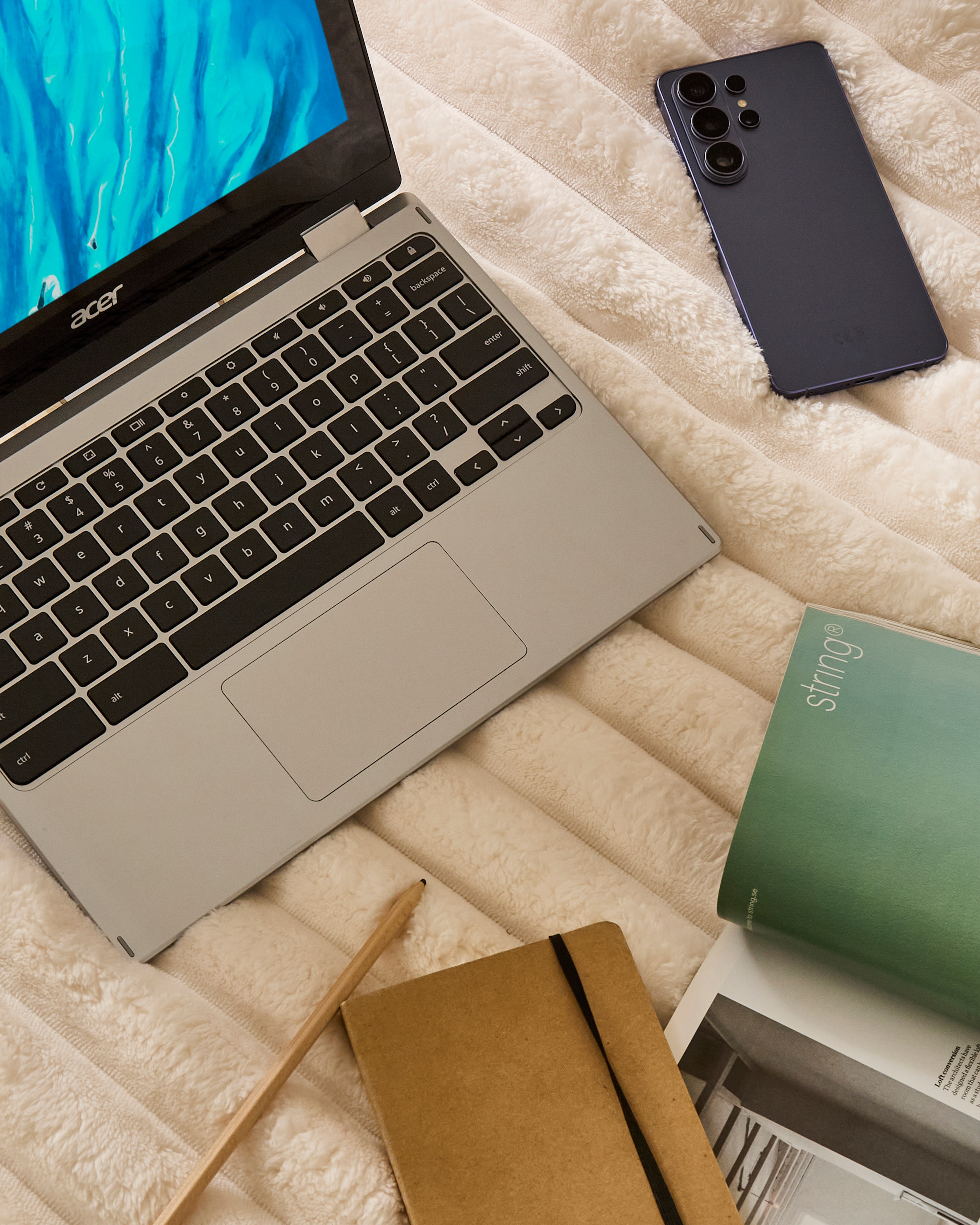 A laptop, a mobile phone and a notepad on a light brown throw blanket.
