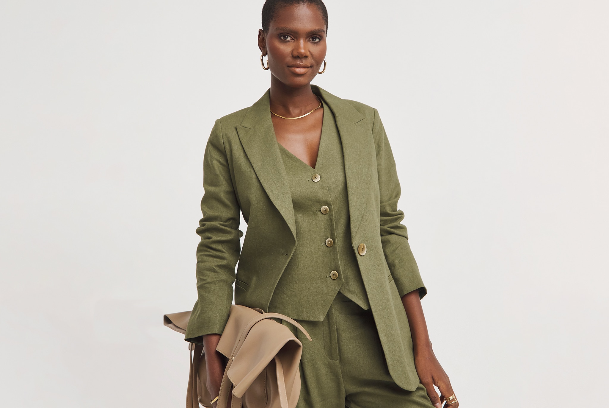 Person wearing an olive-green tailored suit with a buttoned waistcoat and blazer, holding a beige coat