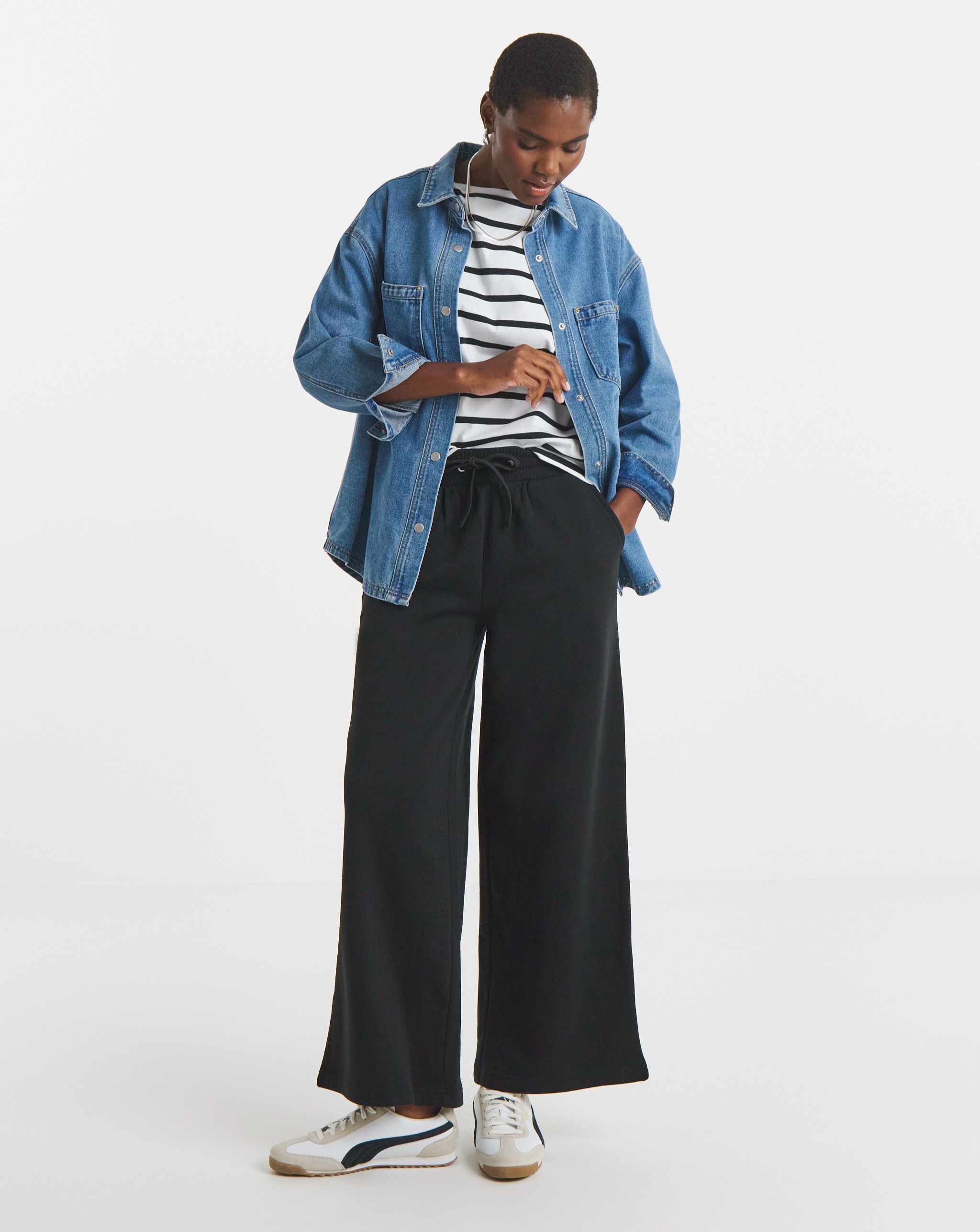 Person wearing a mid-wash denim shirt with a black-and-white striped top, black wide-leg trousers and white trainers.