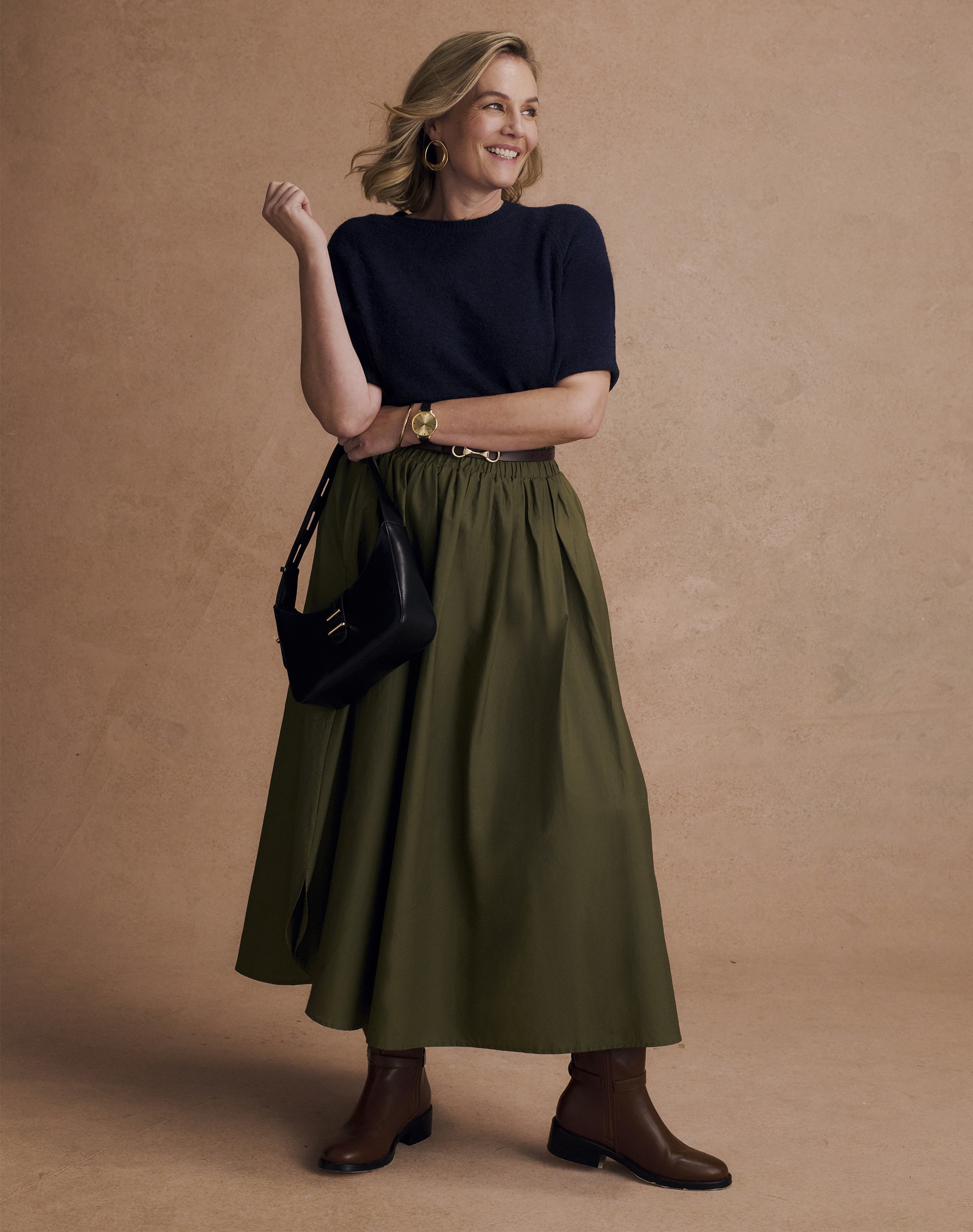 A lady stood in front of a light brown sandy wall wearing a navy short sleeve t-shirt, a green maxi skirt with brown boots.