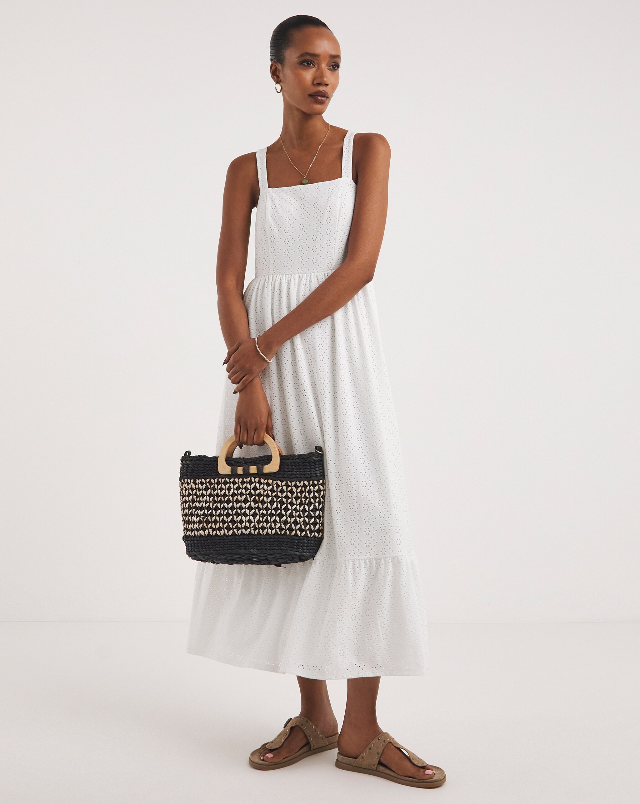 Person wearing a white sleeveless broderie anglaise midi dress with flat sandals, holding a woven black-and-natural tote bag.
